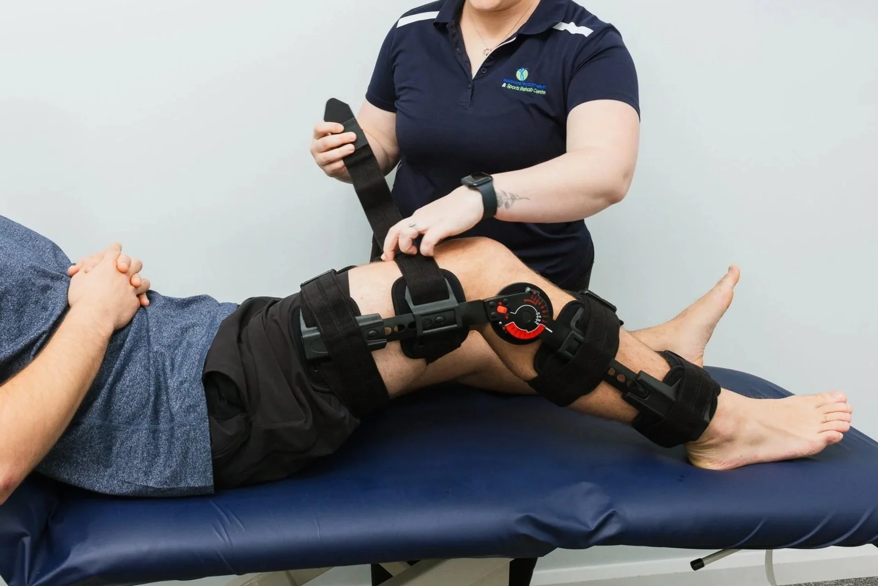 A person lying on a medical table with their leg restrained by a brace and straps, while a healthcare professional adjusts the brace.