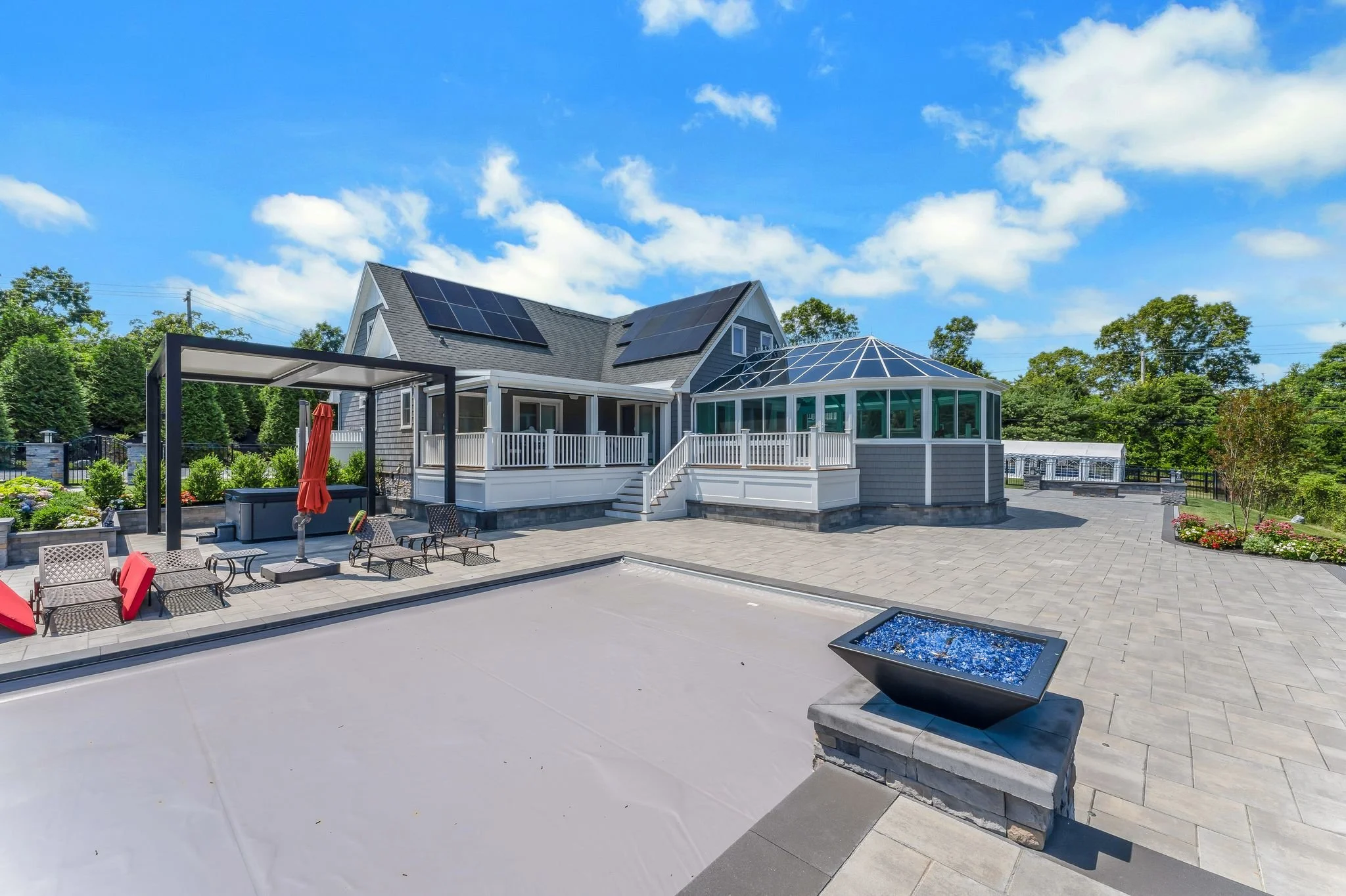 Spacious backyard with a gray house featuring solar panels, a screened porch, and outdoor seating area, surrounded by trees and greenery under a blue sky.