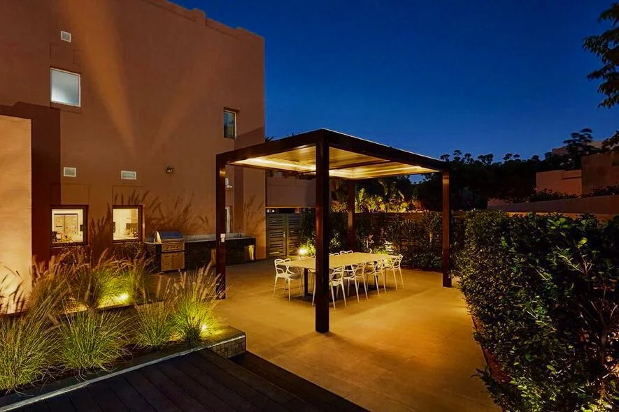 Outdoor patio with illuminated dining table and chairs under a wooden pergola at night, surrounded by plants and a building wall.