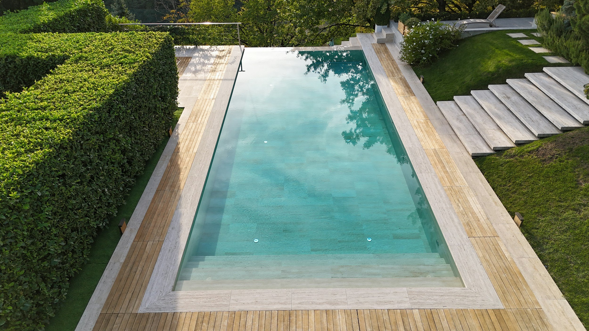 A modern backyard swimming pool with wooden and stone decking, surrounded by green bushes and trees, with outdoor stairs and lounge chairs nearby.