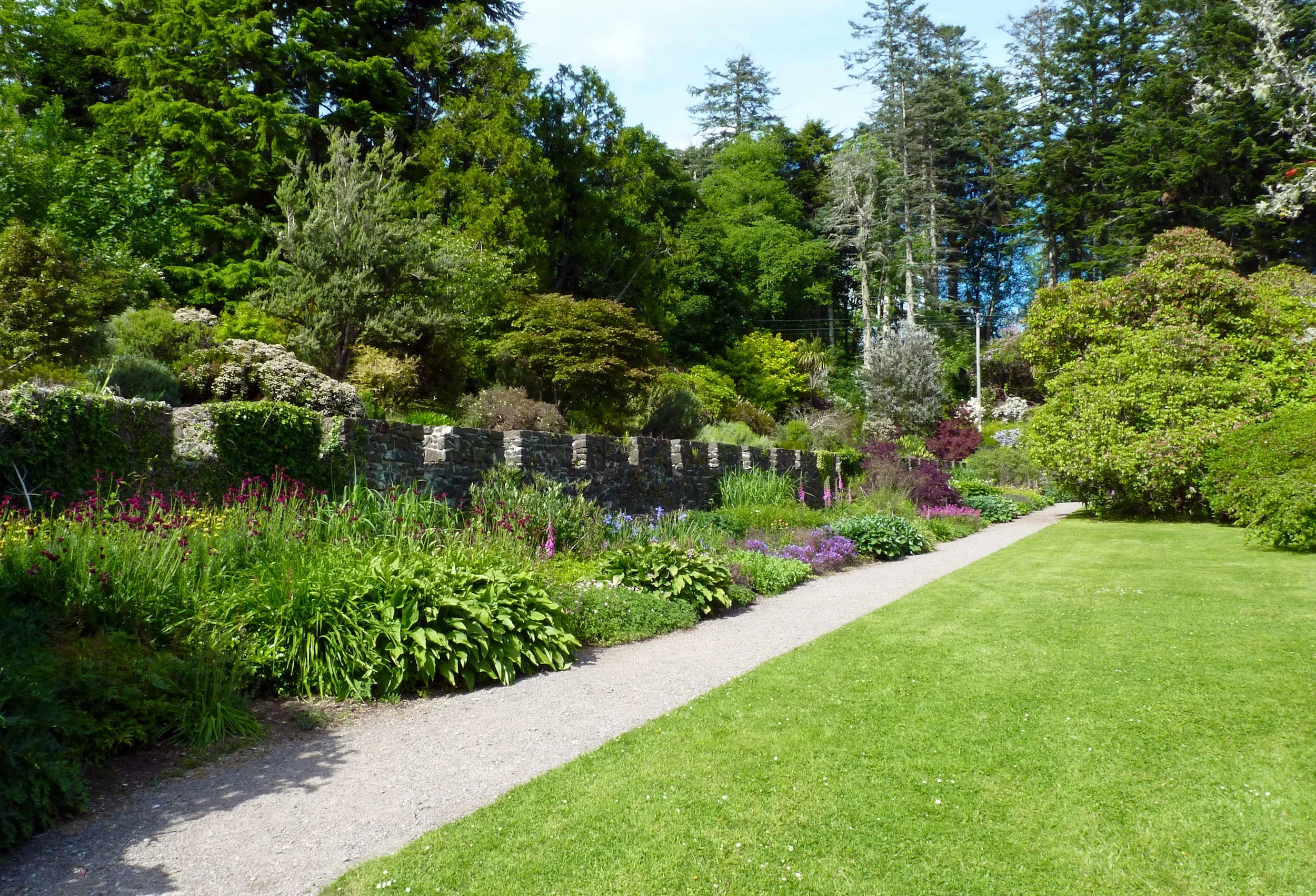A garden with a gravel path running alongside a lush green lawn, surrounded by various flowering plants and trees under a clear sky.