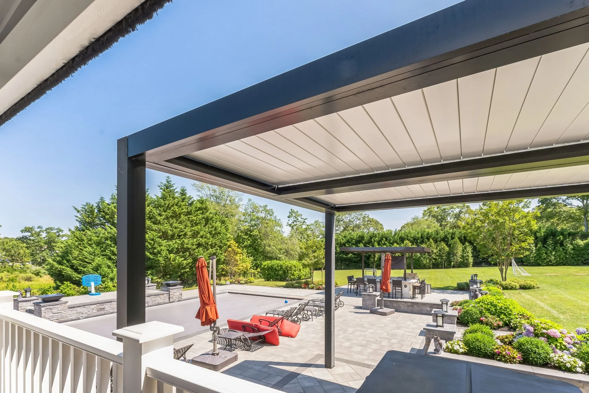 View of an outdoor patio with a shaded area, lounge chairs, umbrellas, a fire pit, barbecue area, and a lawn with trees and a soccer goal in the background.