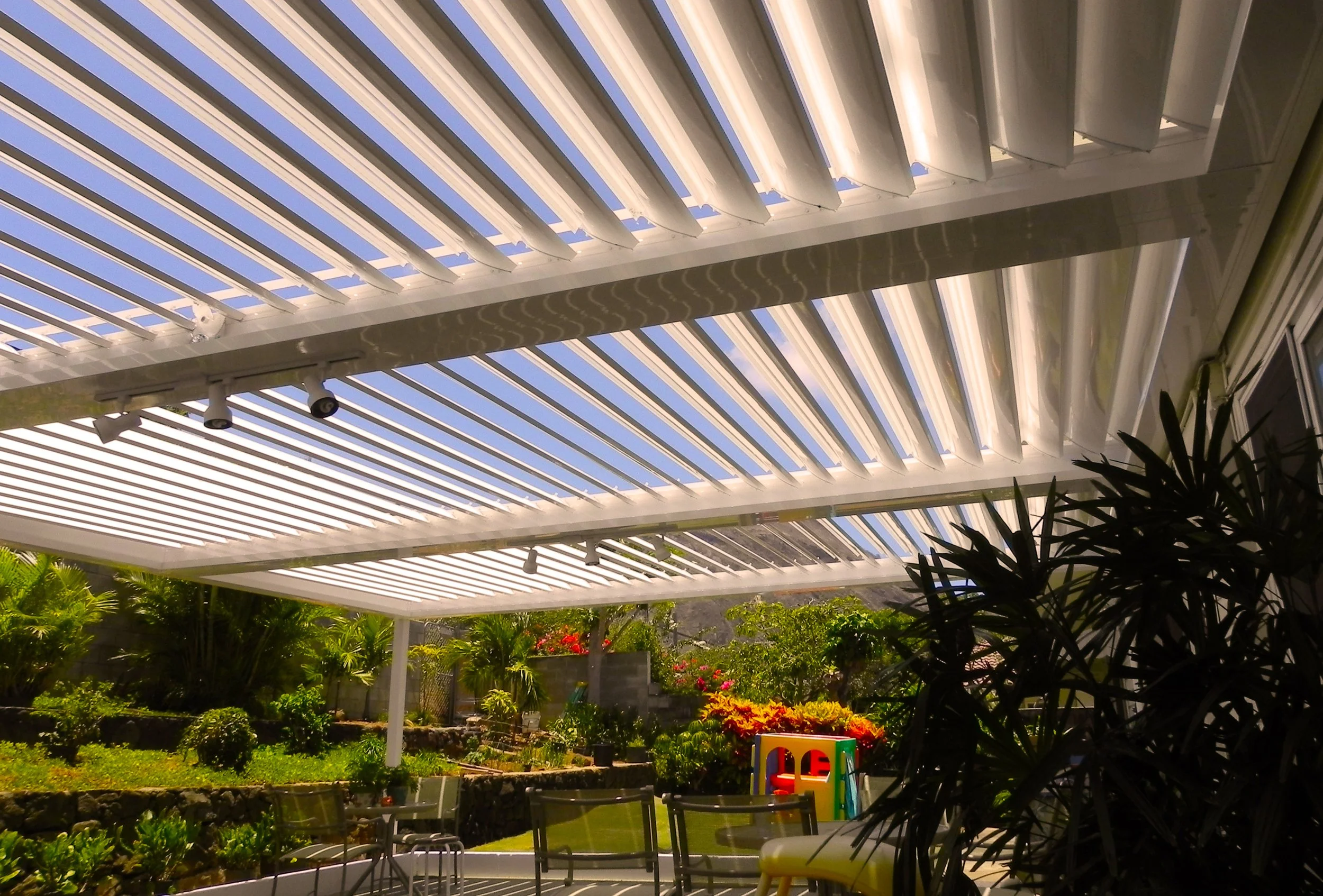 Backyard patio with a white slatted pergola, lush green plants, colorful flowers, and patio furniture.