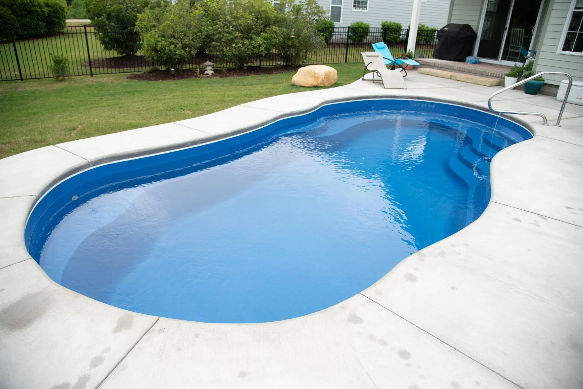 In-ground swimming pool with blue water surrounded by a concrete deck, with a grassy yard and garden with bushes and trees in the background, a black fence, and patio furniture including lounge chairs and a grill.