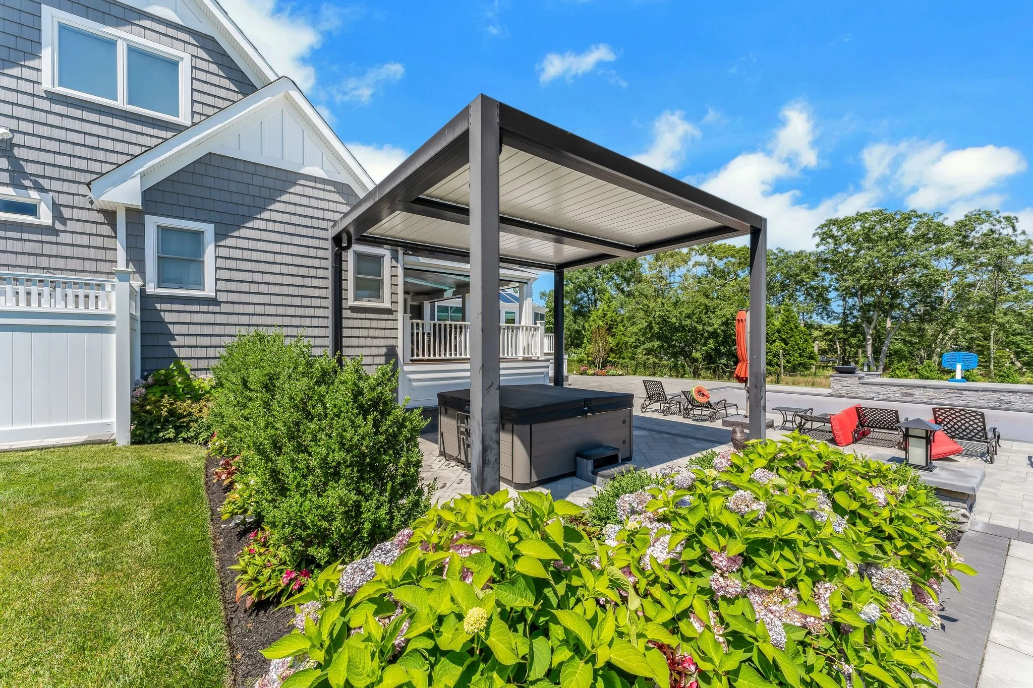 A backyard patio area with a gray house, green bushes, and blooming hydrangeas. There is a covered hot tub and several chairs with red cushions around a fire pit, under a clear blue sky.