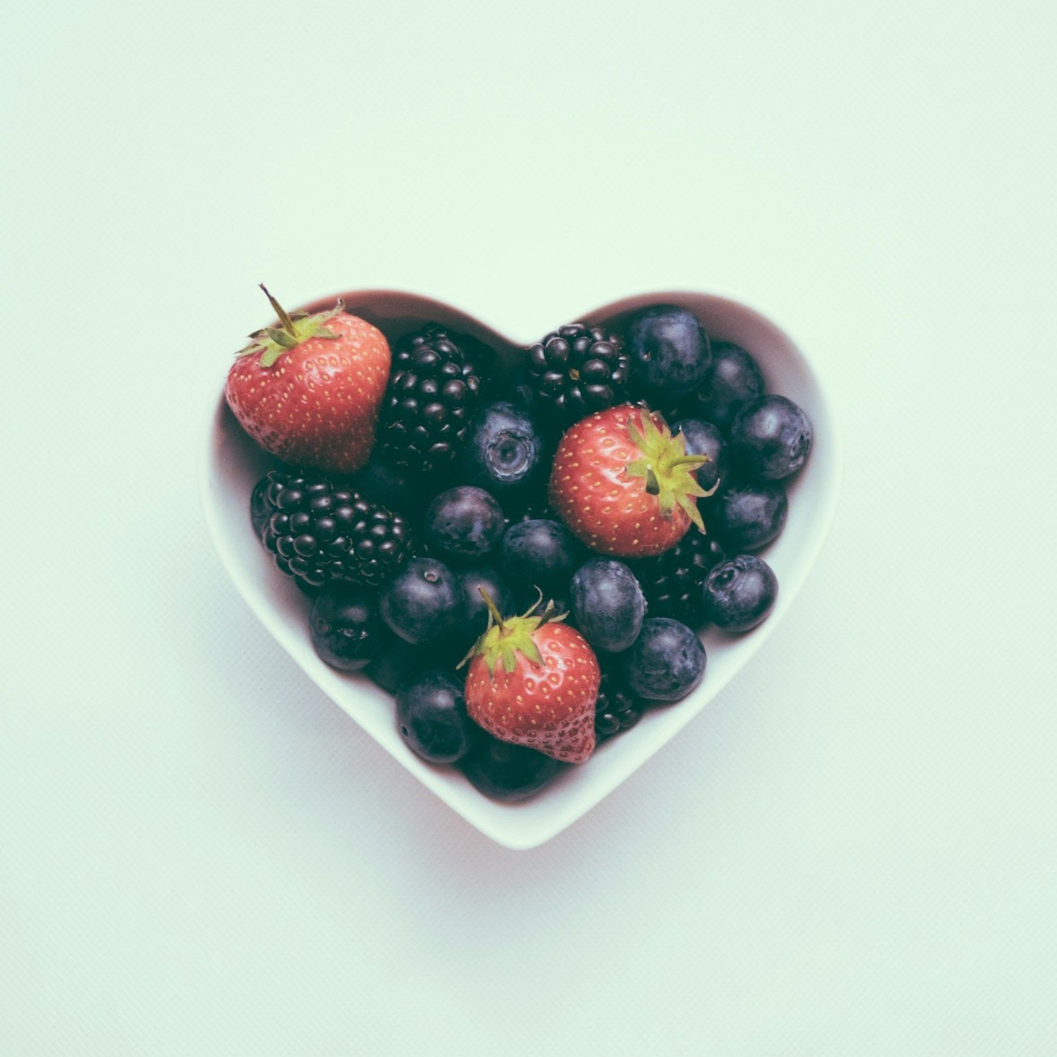 A white heart-shaped bowl filled with strawberries, blackberries, and blueberries on a white surface.