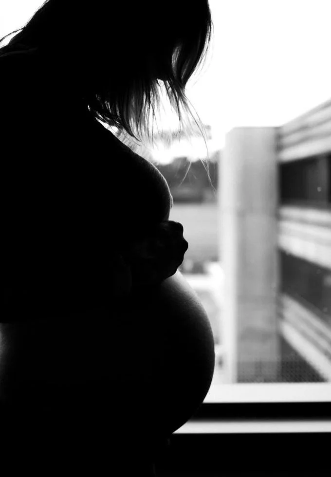 Silhouette of a pregnant woman looking down, standing near a window with an urban exterior view.