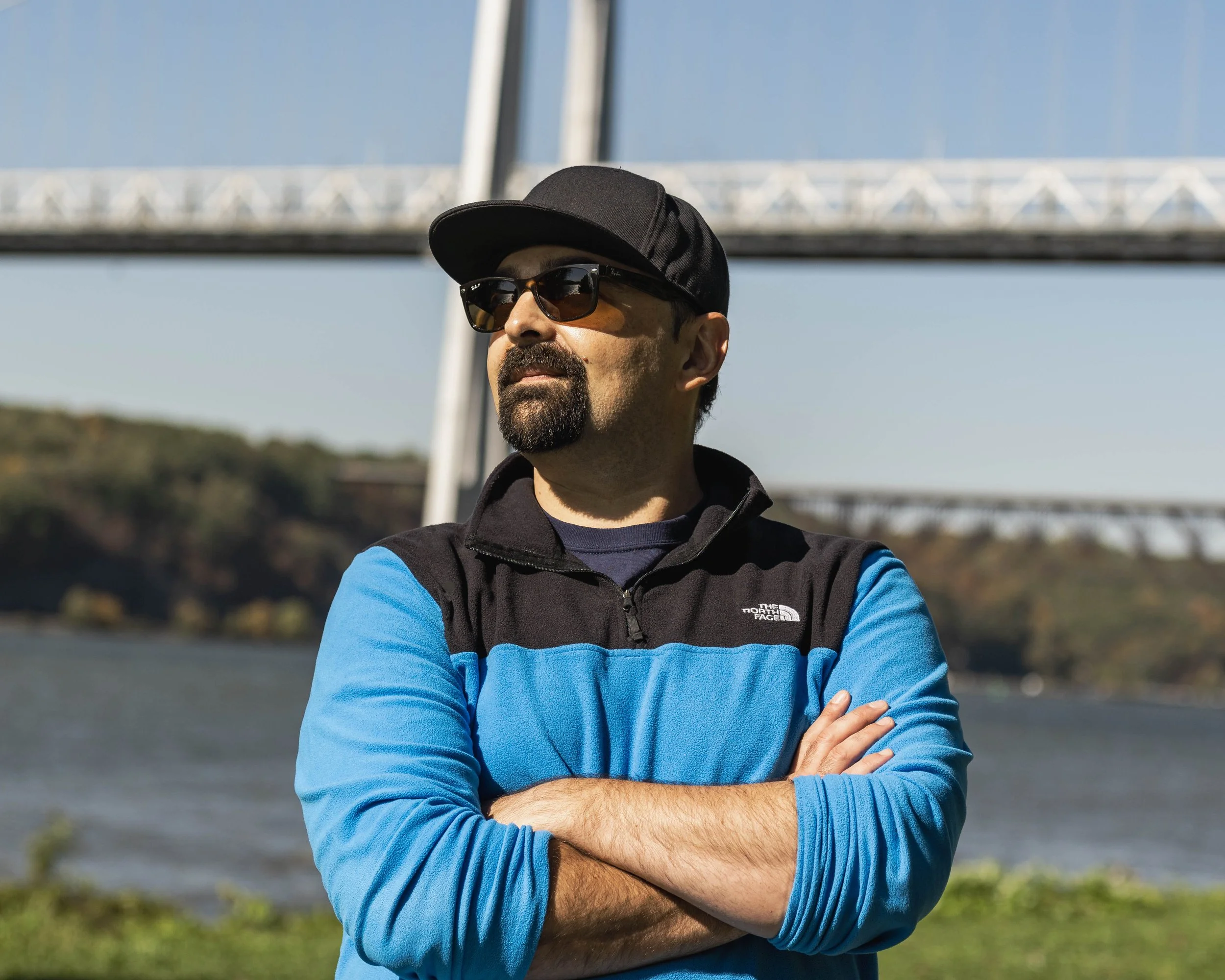 Man wearing a black cap, sunglasses, a blue and black Patagonia jacket, with arms crossed, outdoors near a river with bridges in the background.
