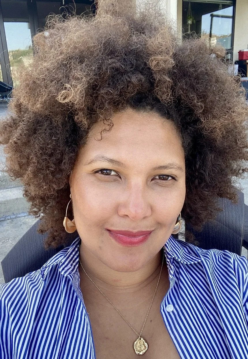 A woman with voluminous curly hair, wearing gold earrings and a gold pendant necklace, smiling outdoors in a blue and white striped shirt.