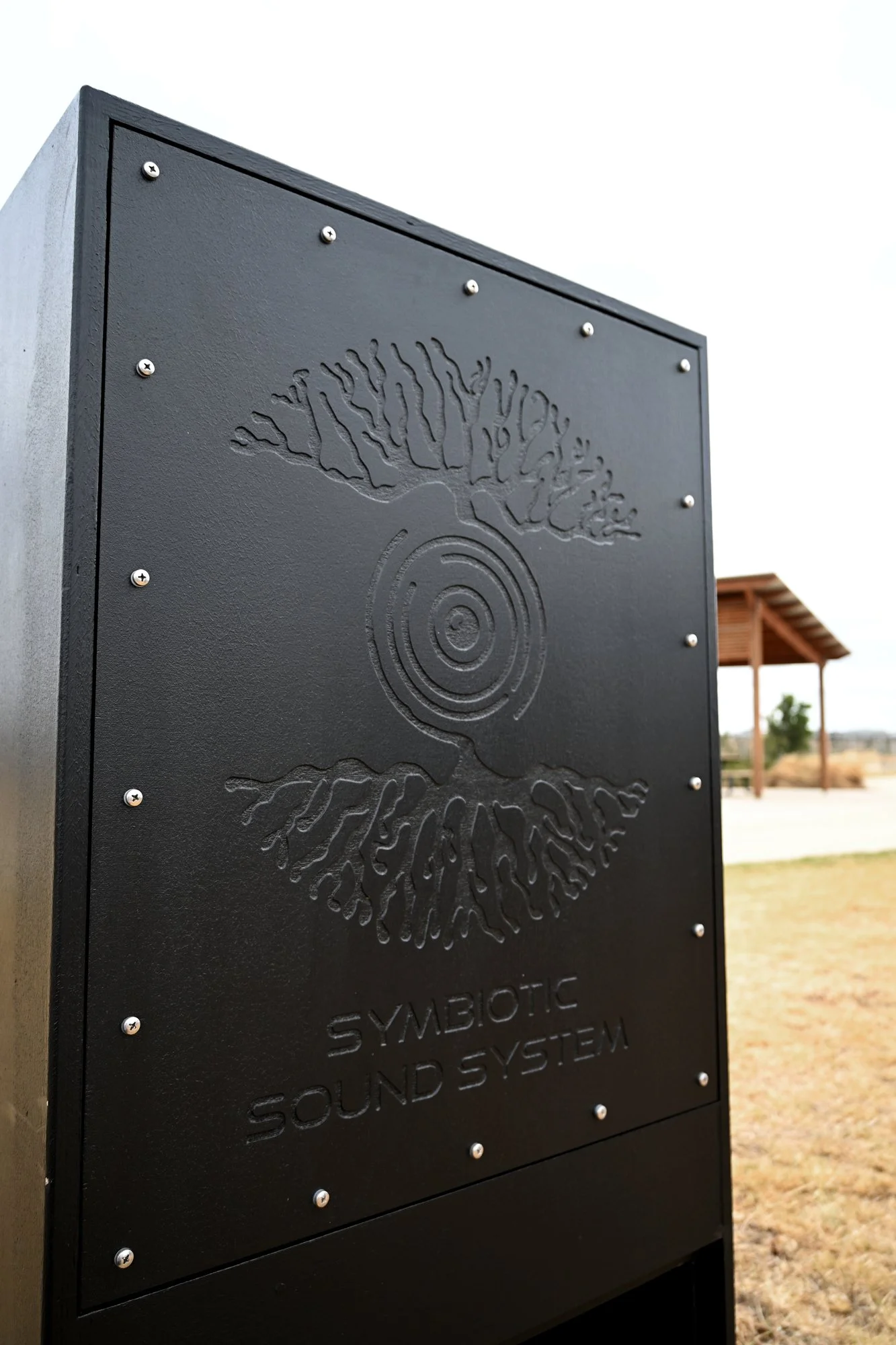 Close-up of a black metal sign with a tree and concentric circle design, labeled 'SYMBIOTIC SOUND SYSTEM', outdoors with a wooden pavilion and open field in the background.