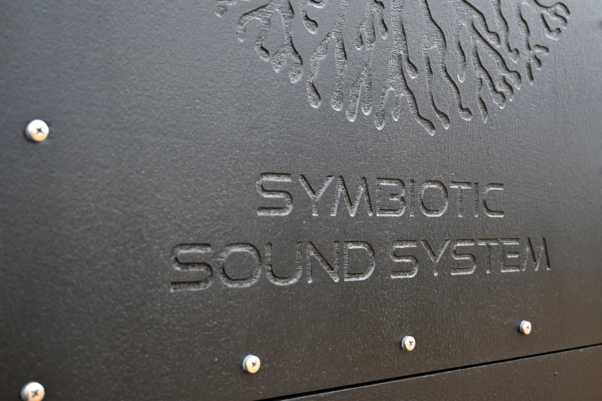 Close-up of a textured black metal surface with the embossed text 'SYMBOTIC SOUND SYSTEM' and a stylized tree logo above it, secured with silver screws.