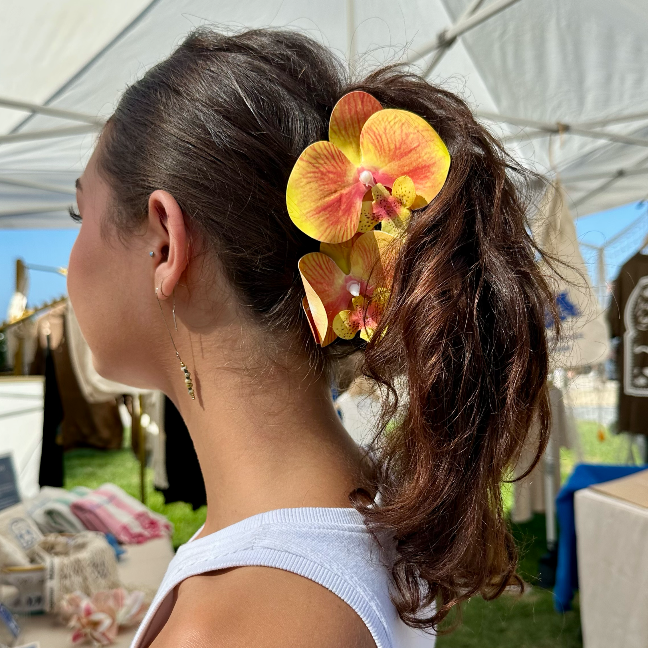 Ponytail styled with an orange orchid hair clip at an outdoor farmers market.