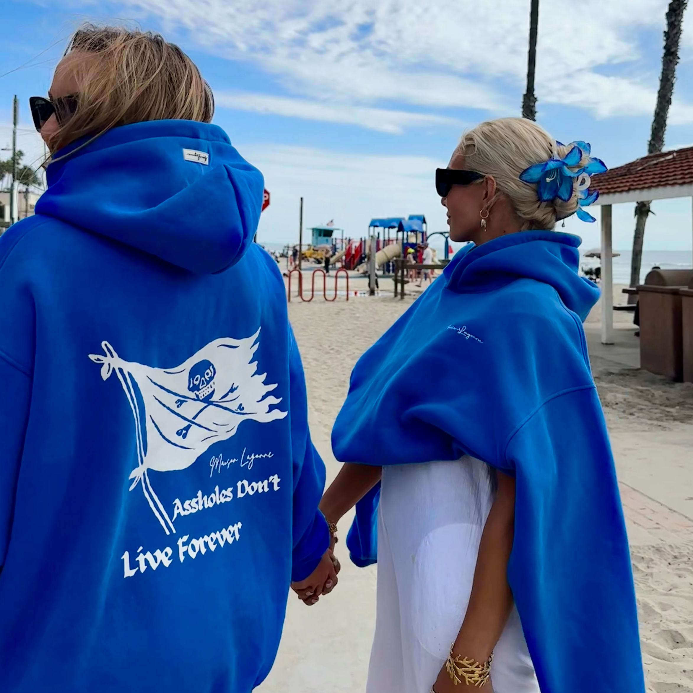 Two people wearing blue hoodies with a white pirate graphic walking by the beach.