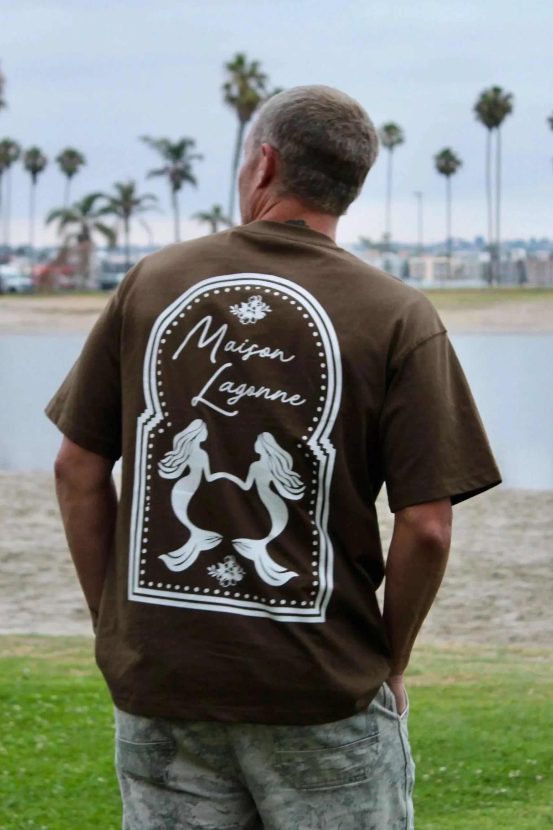 Brown t-shirt featuring the Maison Lagonne twin mermaid logo worn by the water.