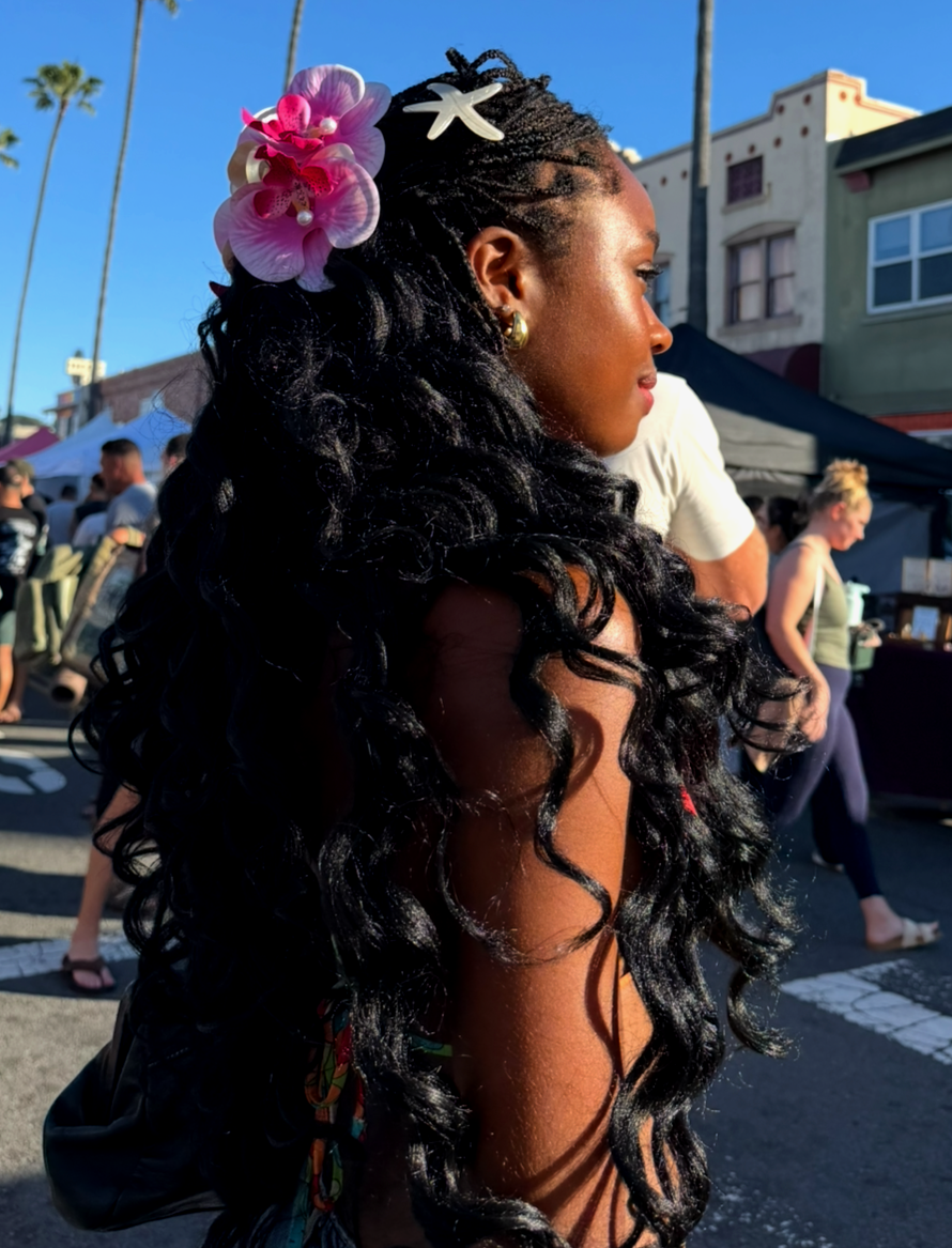 Black wavy hair styled with a pink orchid  hair clip at an outdoor market.
