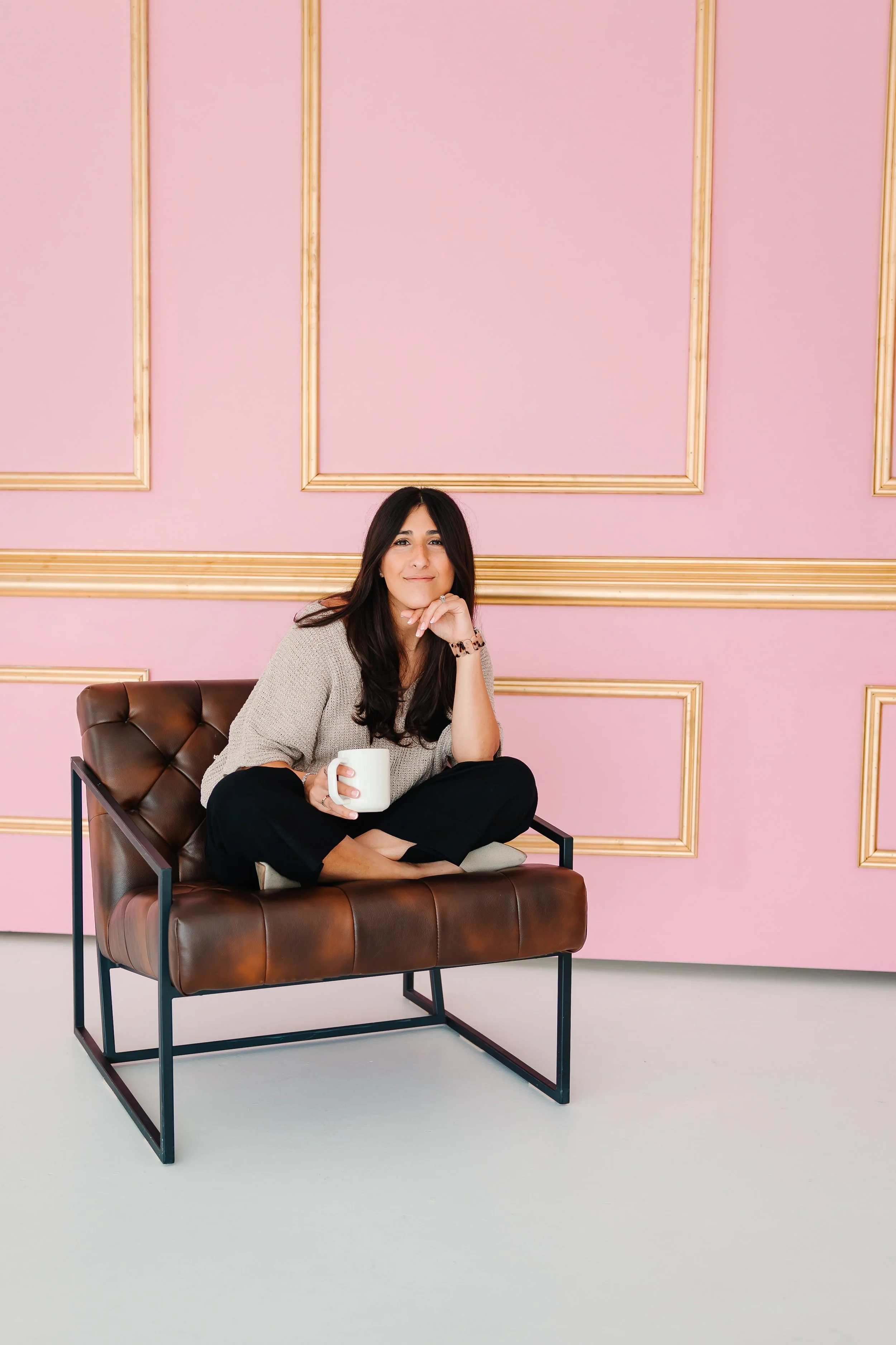Woman with long dark hair sitting cross-legged on a brown leather chair against a pink wall with gold decorative trim, holding a white mug and smiling at the camera.