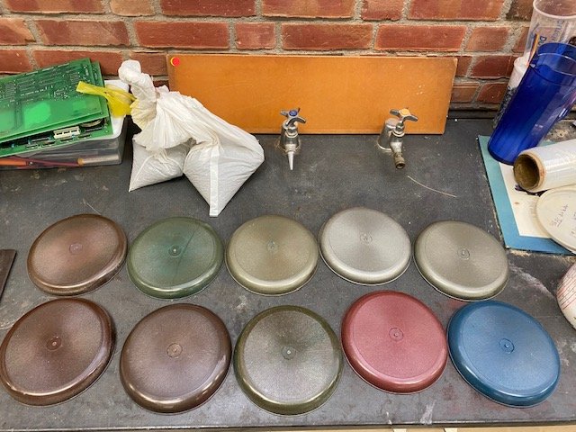 A row of ten colored lids on a work surface with a brick wall in the background, along with various tools, a blue water bottle, and miscellaneous items.