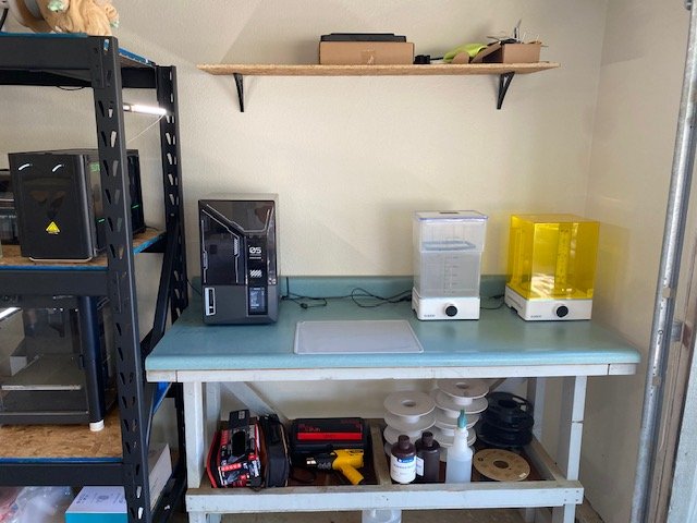 A workspace with a metal shelf on the left, a blue workbench in the center with various equipment, bottles, and tools underneath, and a wooden shelf above with items stored on it.