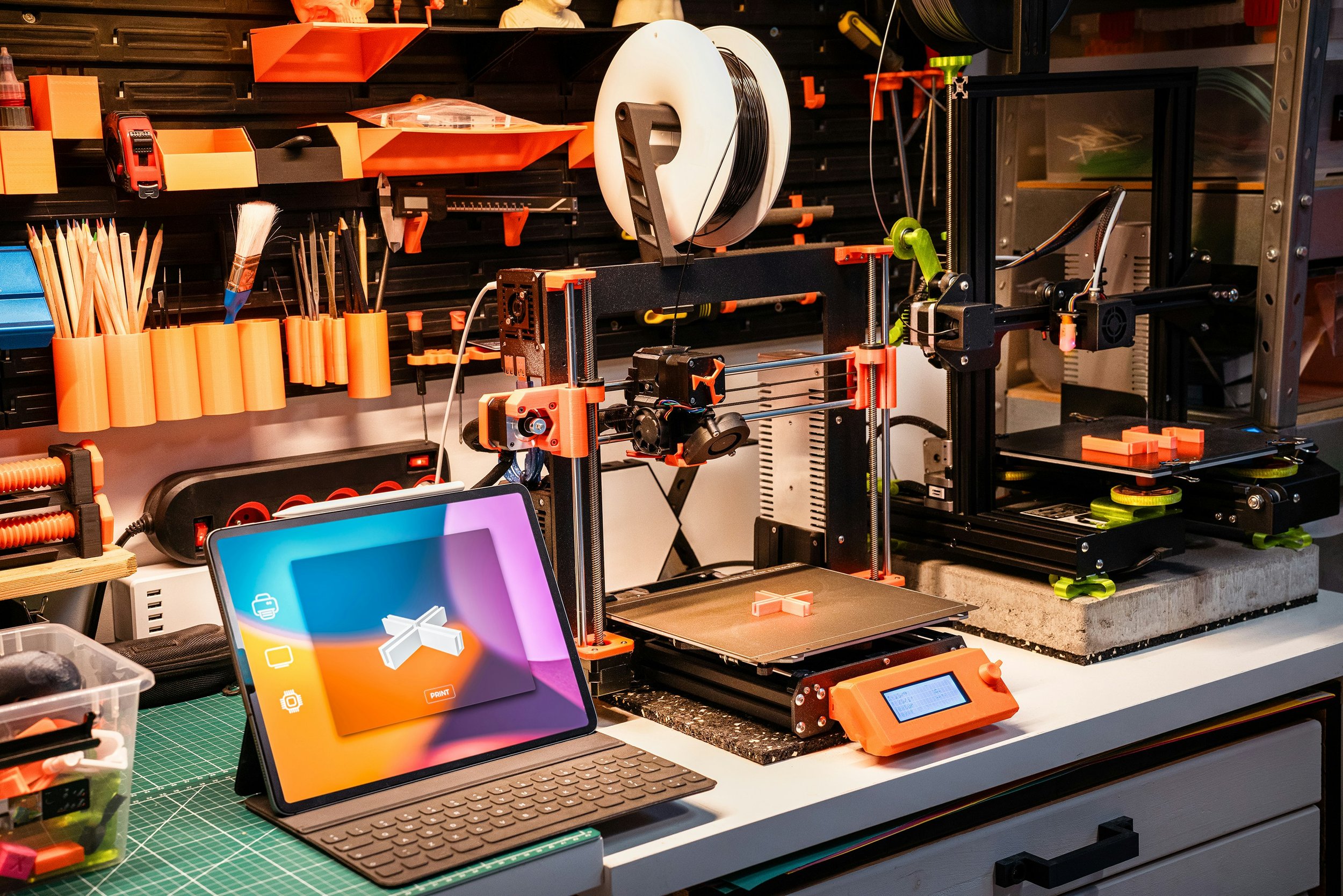 A workspace with 3D printers, tools, and a tablet on a green cutting mat for designing and manufacturing 3D-printed objects.