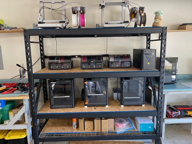 A metal shelving unit with three shelves in a workshop. The top shelf has 3 3D printers with filament spools. The middle shelf has 3 printer enclosures. The bottom shelf has boxes, a glue bottle, and plastic filament spools. To the left, a workbench