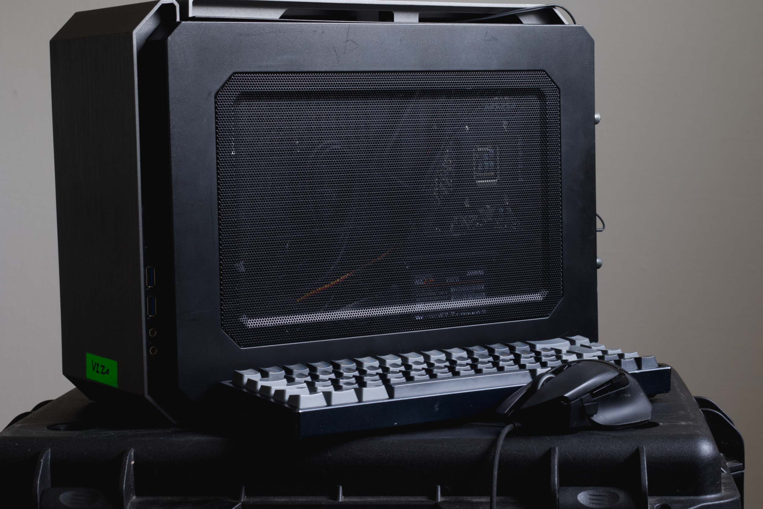 A black computer case with a mesh front panel, a white keyboard, and a black mouse, placed on a black surface against a plain wall background.