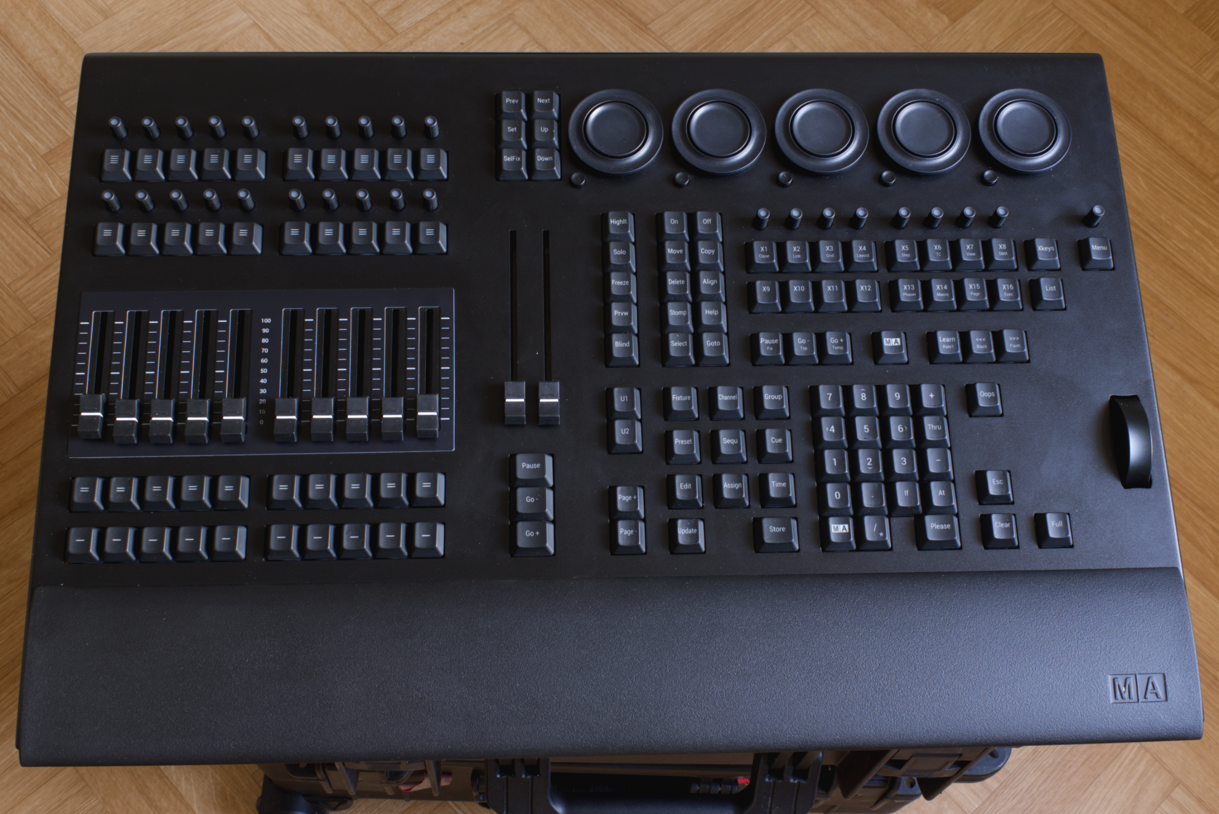 Overhead view of a large professional audio mixing console with multiple sliders, knobs, buttons, and a mouse on a wooden floor.