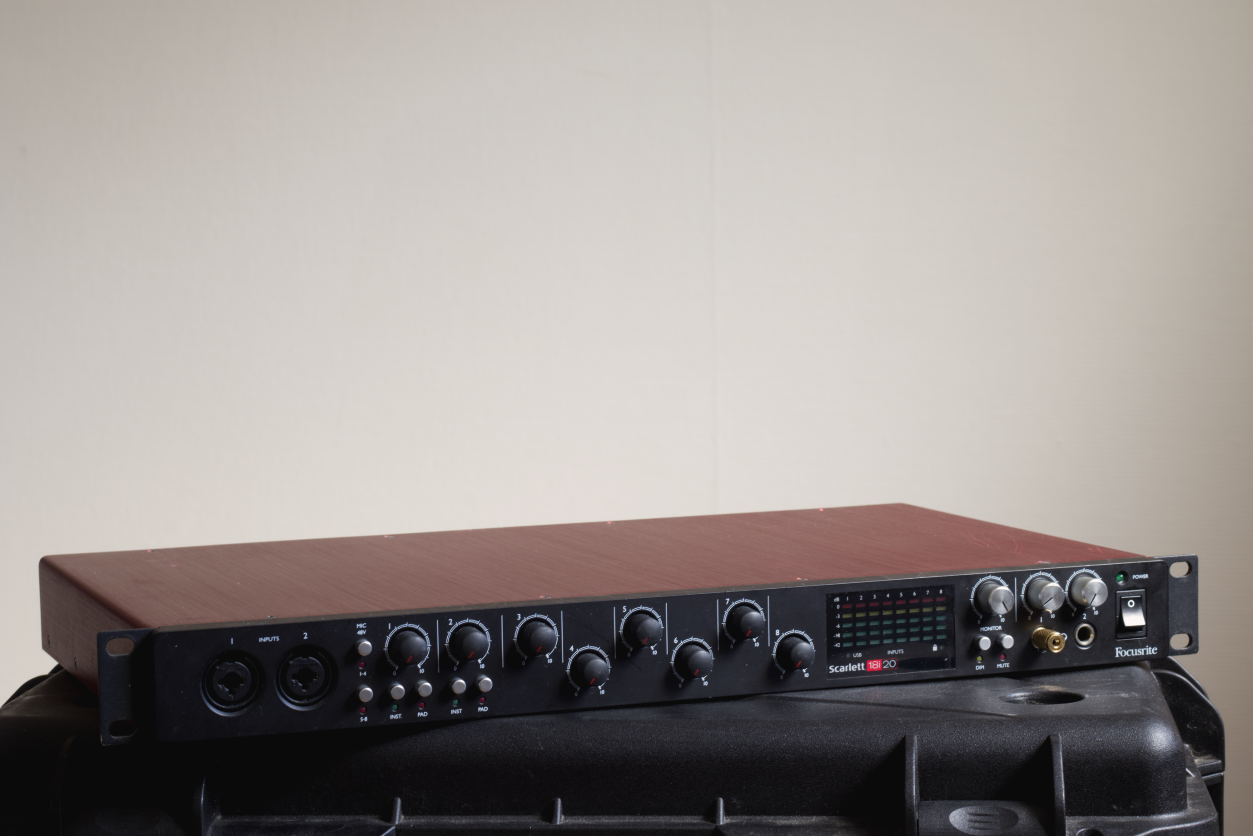Audio mixer with knobs, inputs, and display, placed on a black case against a plain wall.