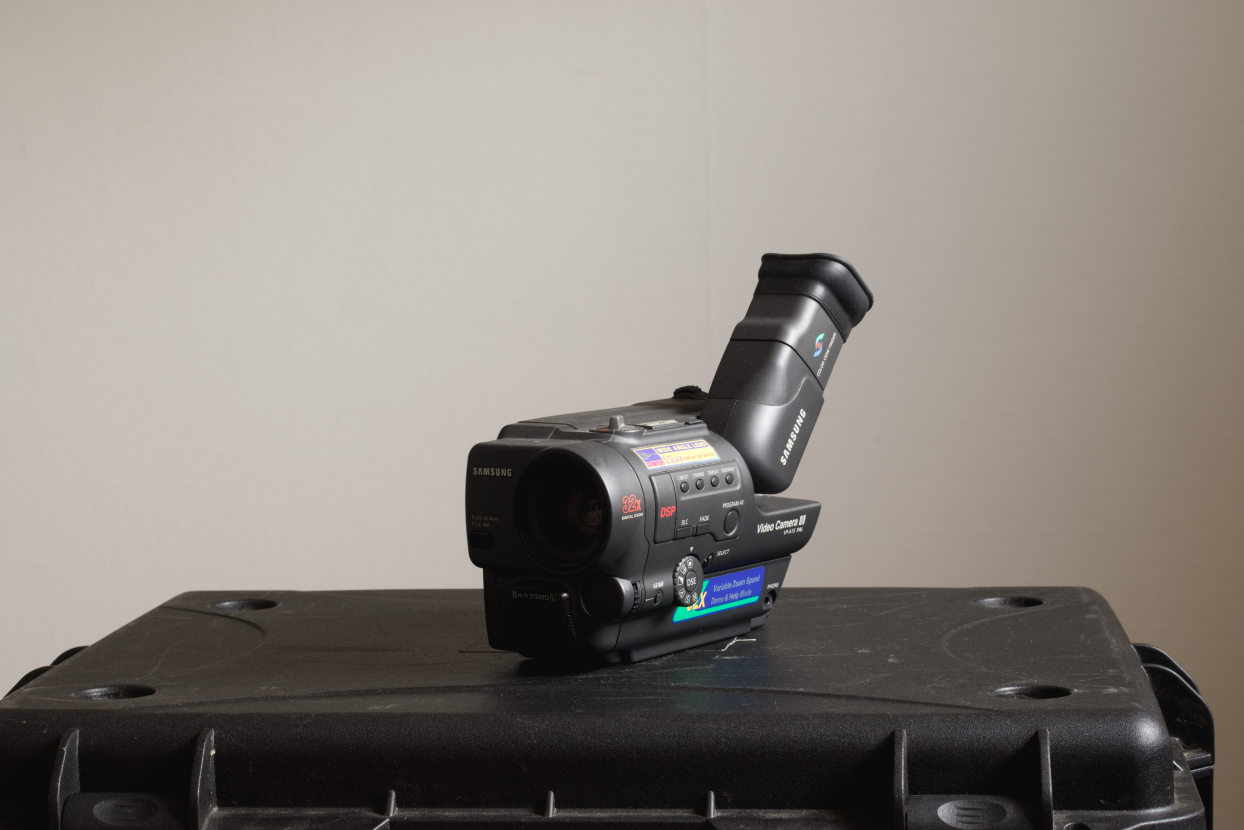 A black Samsung camcorder with a flip-out screen, placed on top of a black plastic case against a plain beige wall.