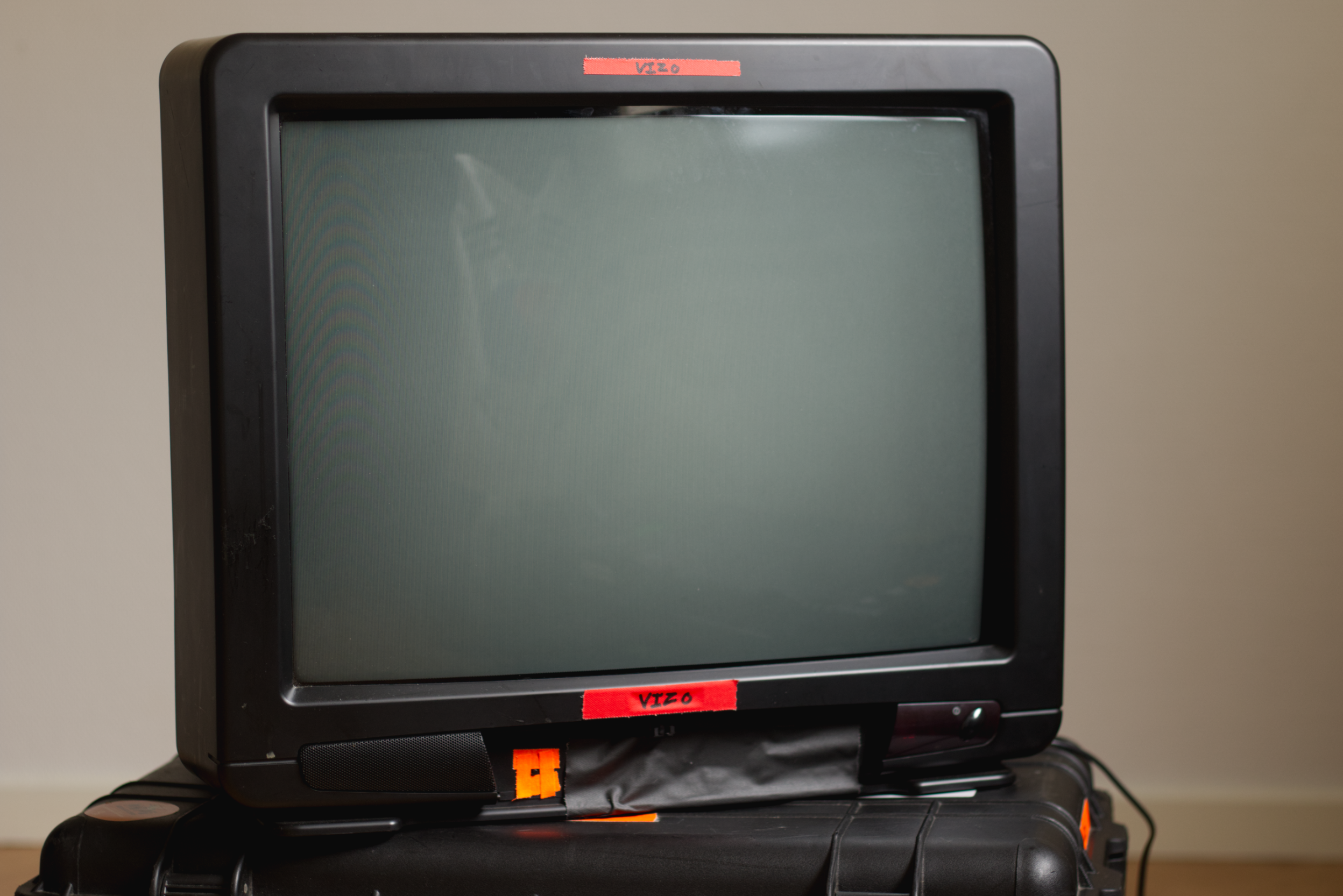 An old black television set with a blank screen, placed on top of a black suitcase, against a plain wall background.
