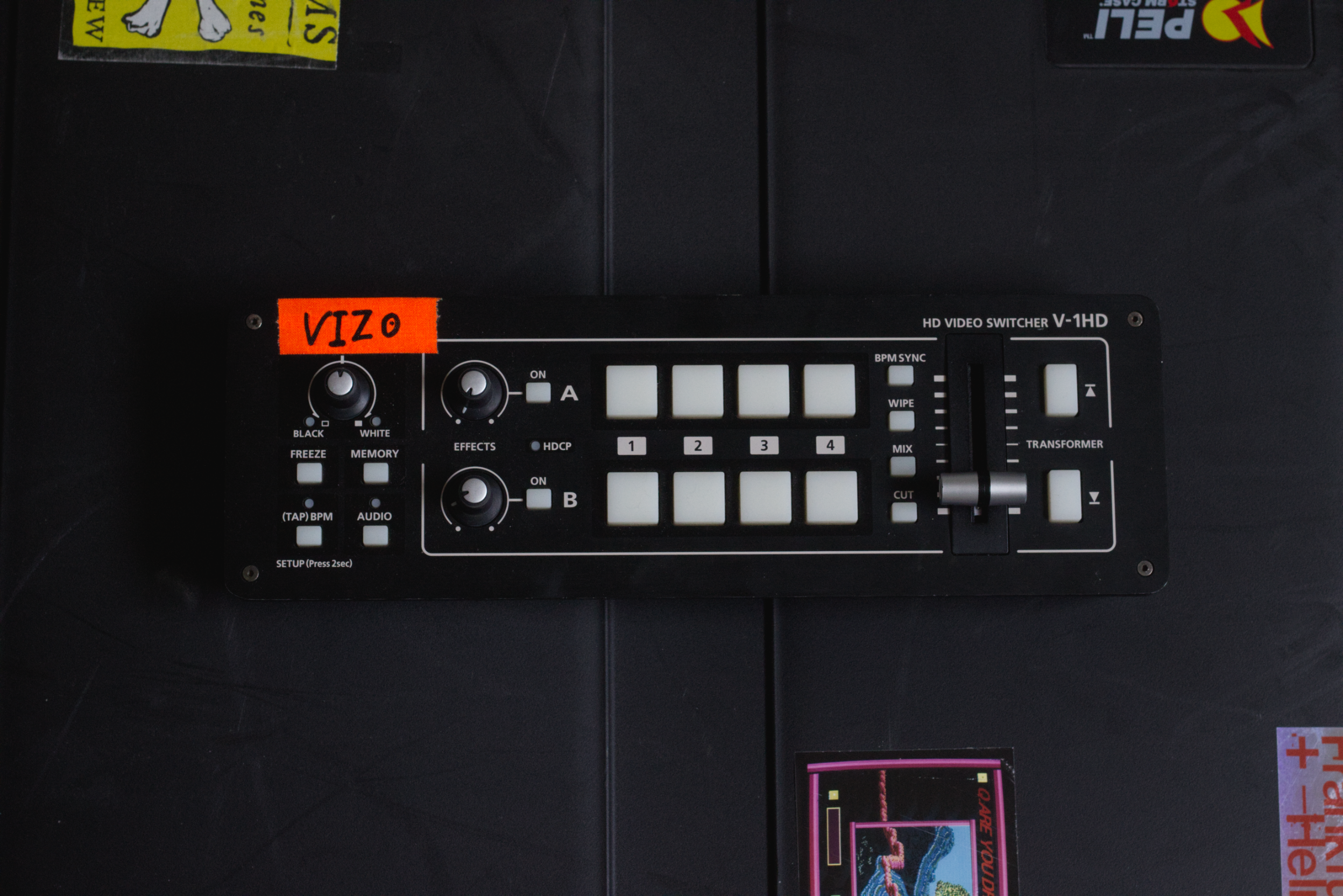 A black Roland V-1HD HD video switcher with white buttons and knobs, mounted on a black surface, with some small stickers partially visible around it.