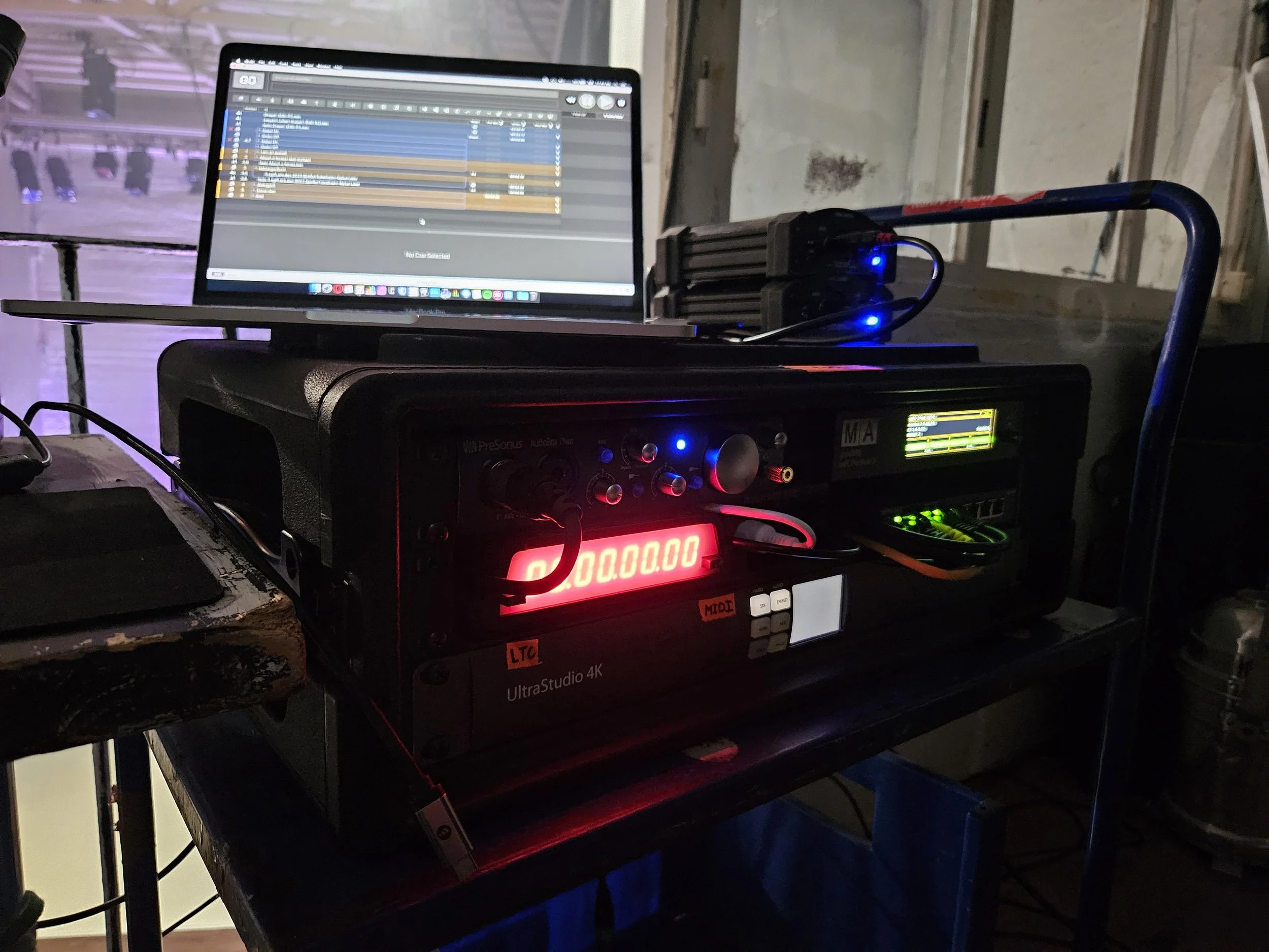 A music production setup featuring a computer monitor displaying a digital audio workstation, a Pro Suite audio interface, and an UltraStudio 4K capture device on a black rack, with various cables connected in a dimly lit environment.