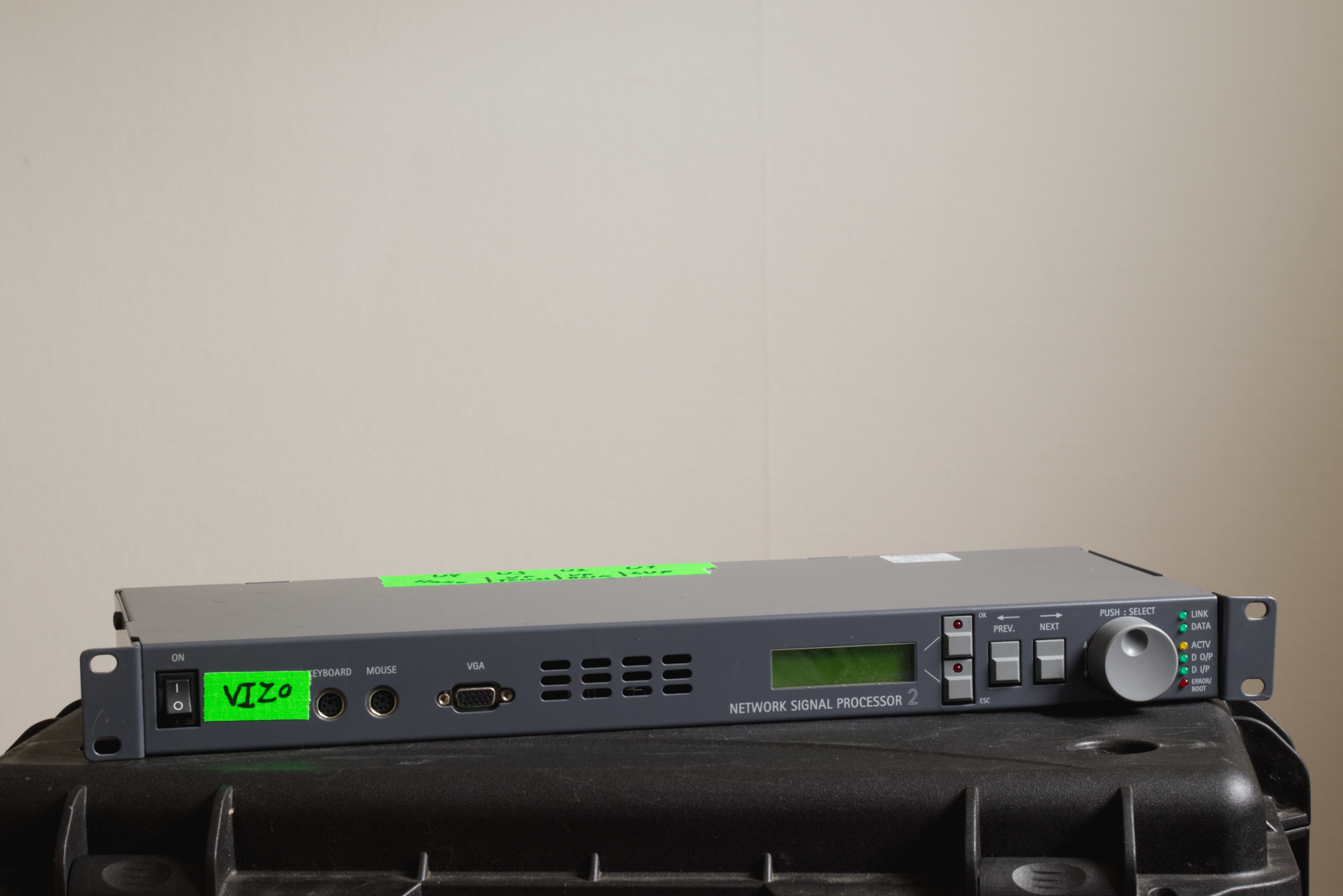A network signal processor rack-mount unit with a green label reading 'VIZO', an LCD screen, various buttons, and indicator lights, resting on a black surface against a plain wall background.
