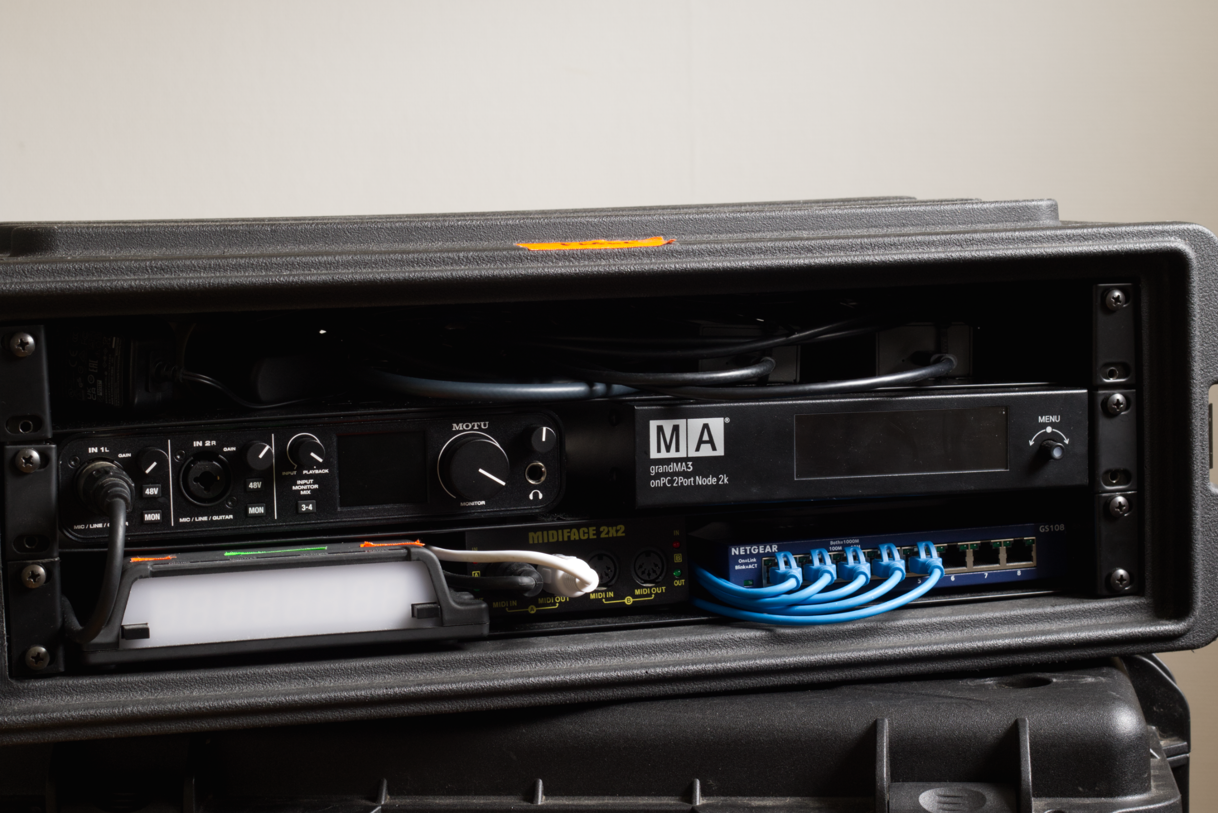 Audio equipment rack with various electronic devices, including a network switch, MIDI interface, and audio interface, housed in a black case.