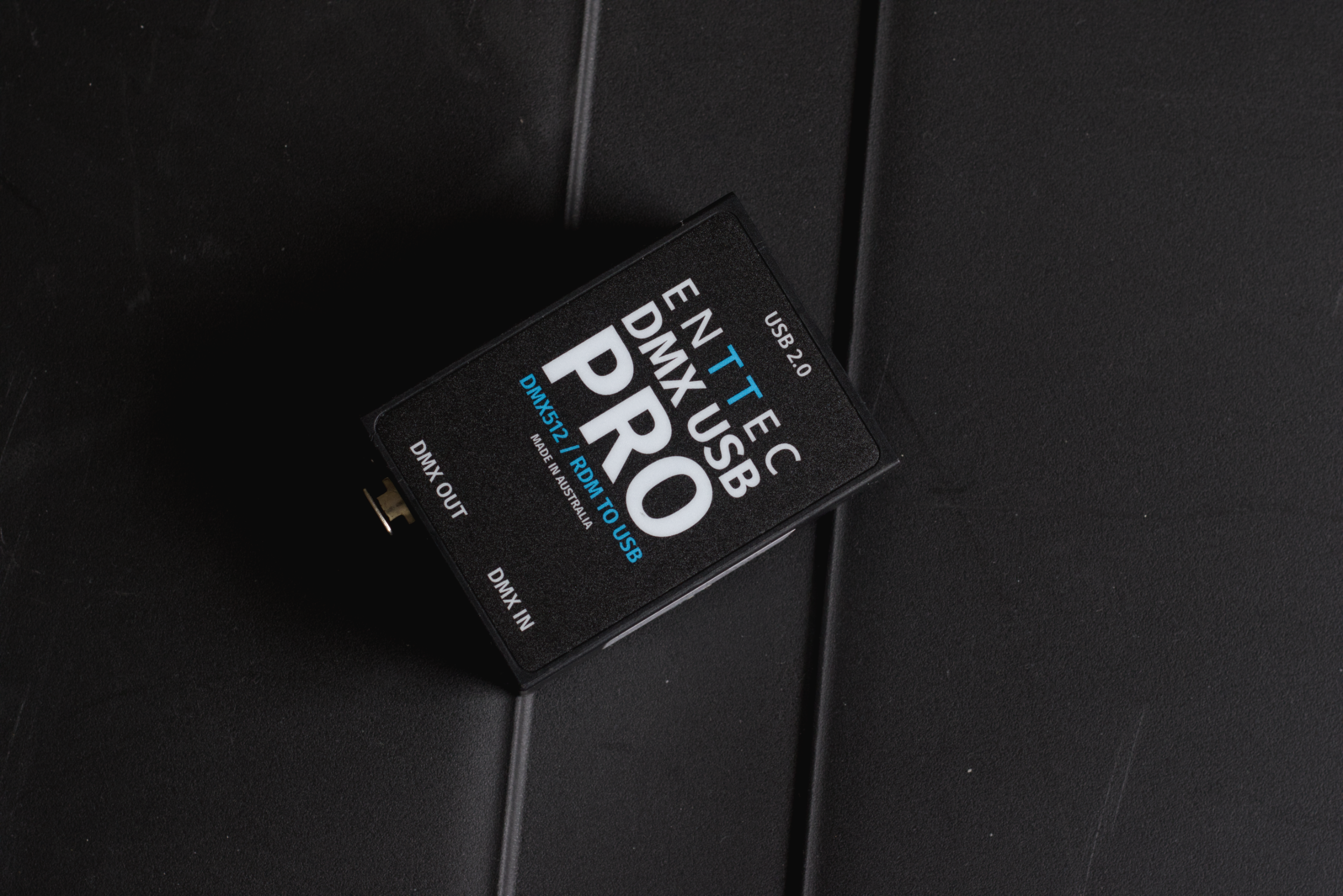 A black electronic device labeled TEC DMX USB PRO with USB 2.0 and DMX IN/OUT ports, placed on a black surface.