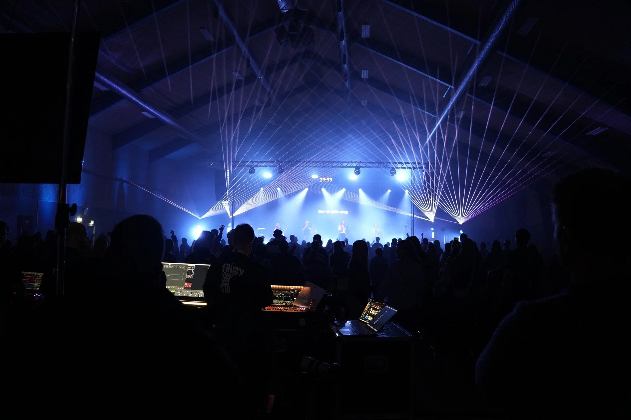 Dark concert venue with silhouettes of audience facing a brightly lit stage with laser lights.