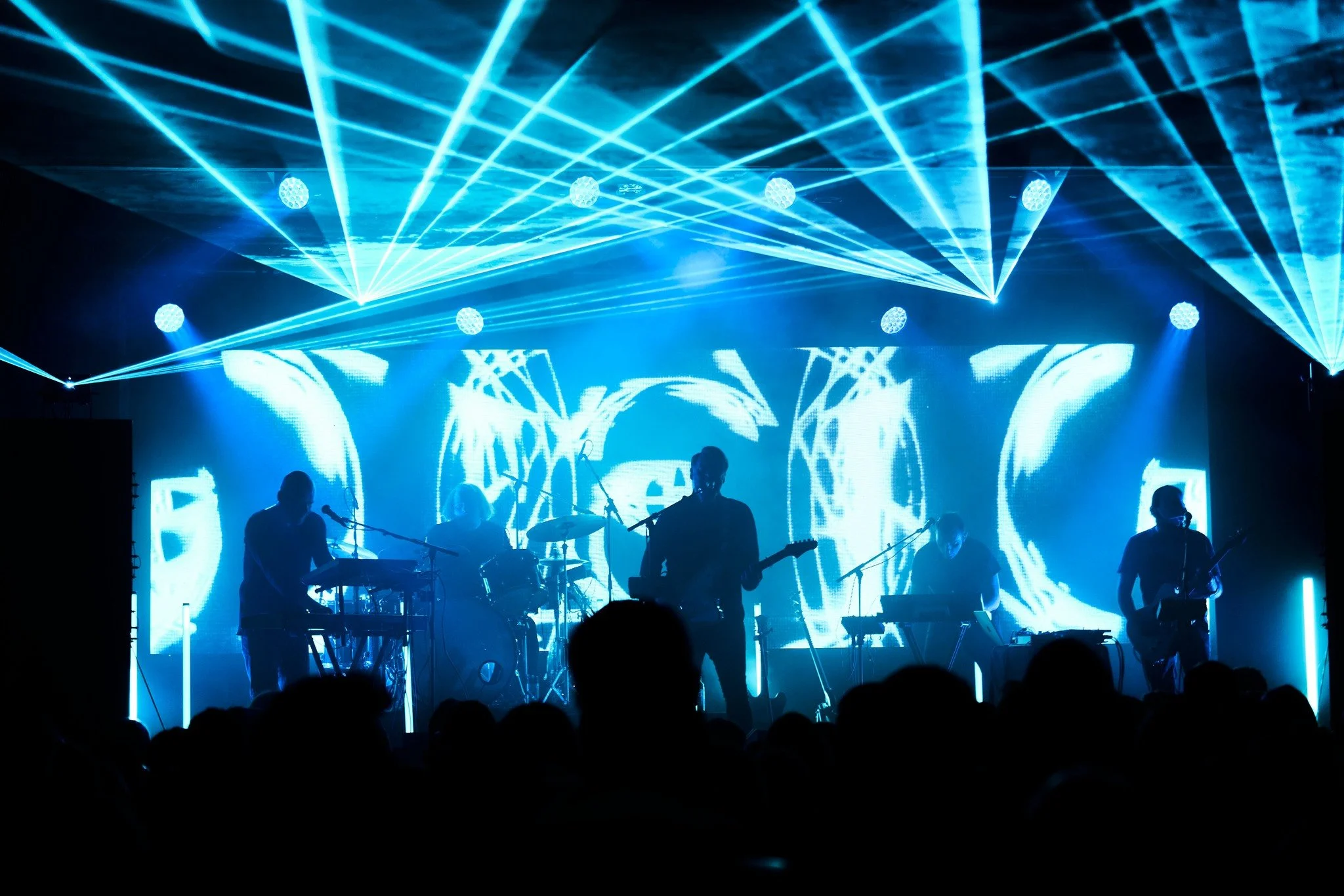 A band performing on stage with blue lights and laser beams, silhouettes of musicians in front of a digital backdrop, audience in foreground.