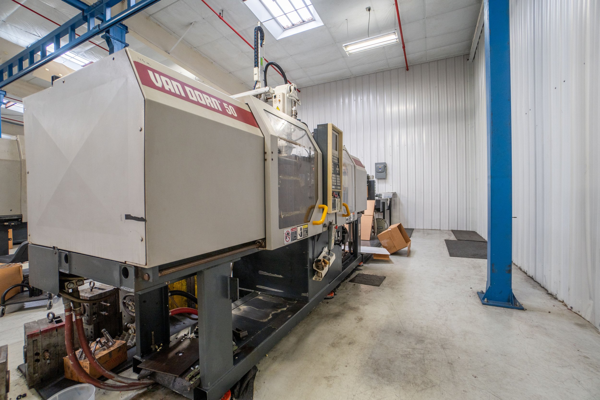 Industrial manufacturing equipment in a warehouse with white walls and a concrete floor, with some cardboard boxes and floor mats nearby.