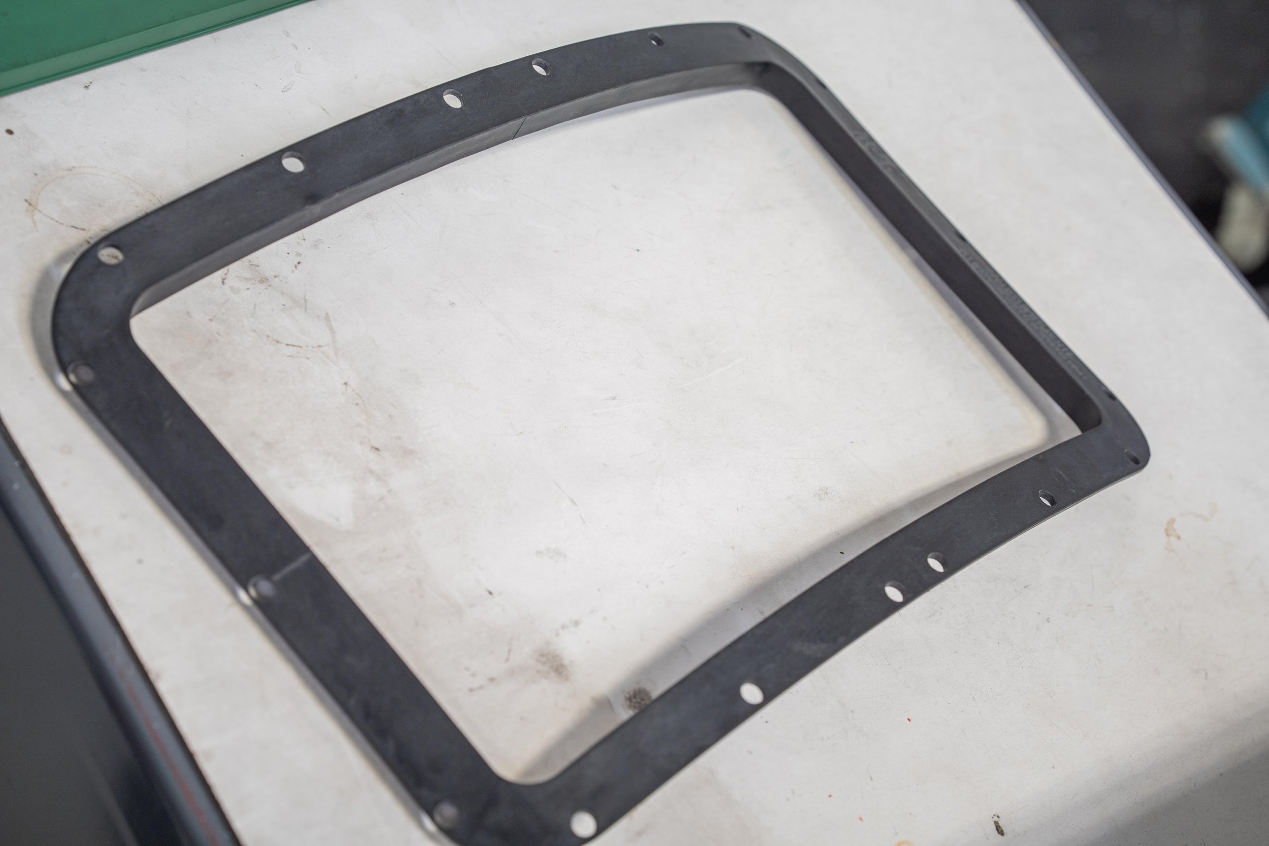 A black rectangular gasket with multiple holes laid on a white work surface.
