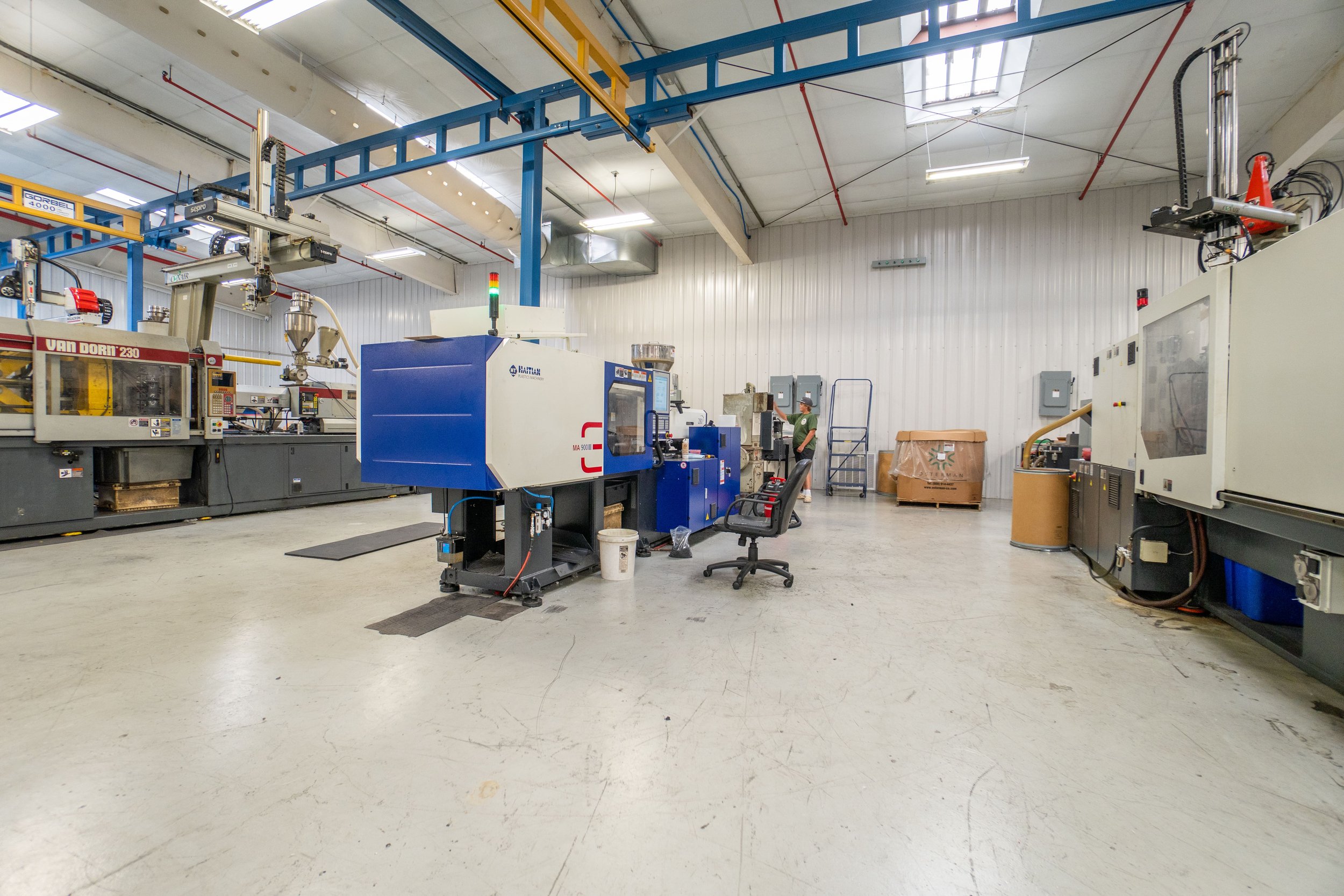 A manufacturing workshop with various industrial machines, including CNC and automated equipment, and workers operating machinery in a spacious, well-lit environment with a high ceiling, white walls, and a yellow overhead crane.
