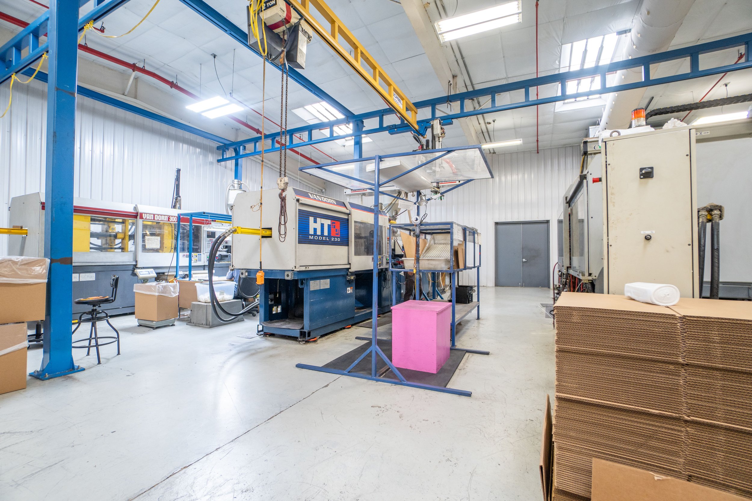 Industrial manufacturing or printing facility with large machines, conveyor belts, cardboard boxes, and stacks of flattened cardboard packaging.