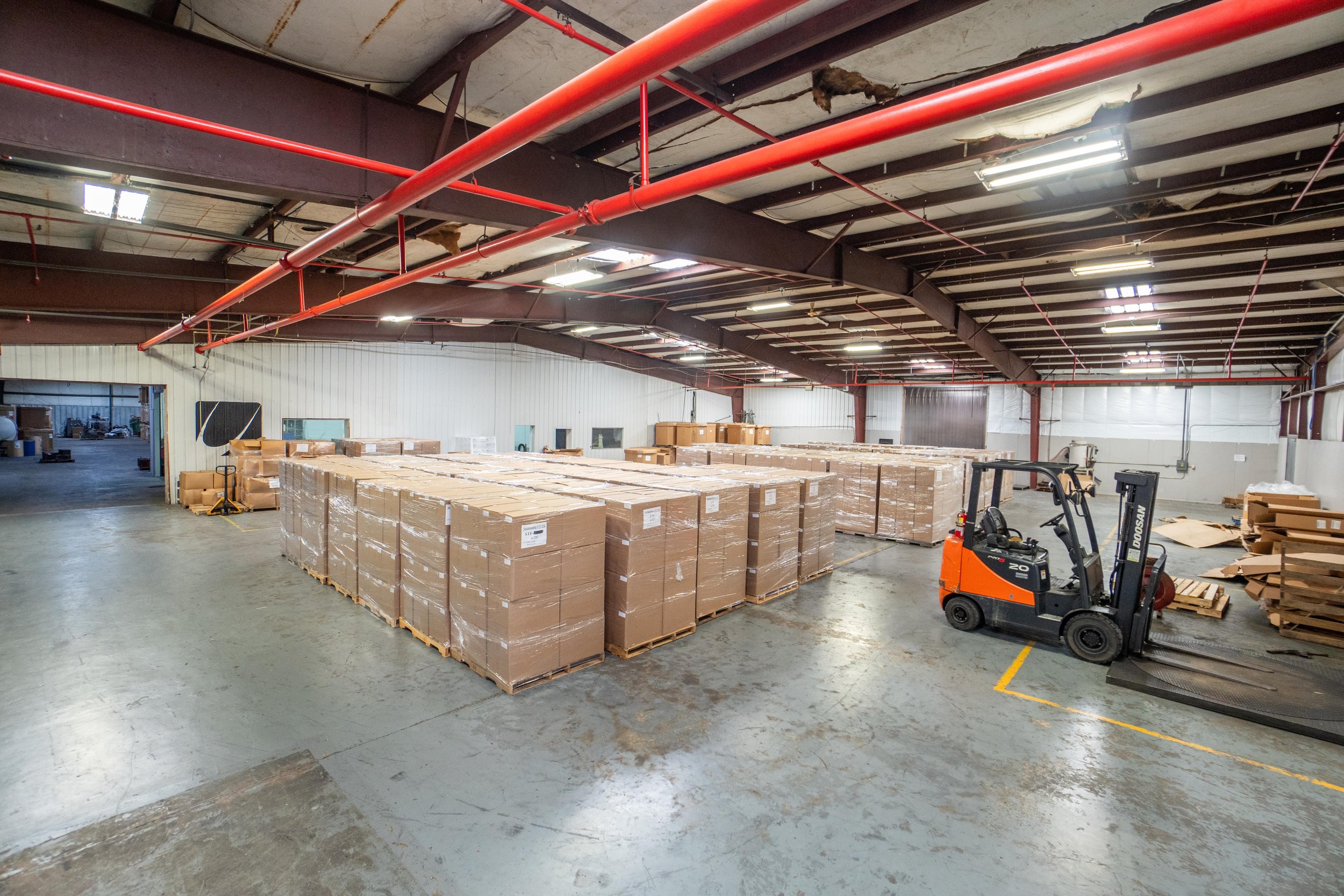 Warehouse interior with stacked cardboard boxes on pallets, a forklift, and unfinished ceiling with visible beams and pipes.