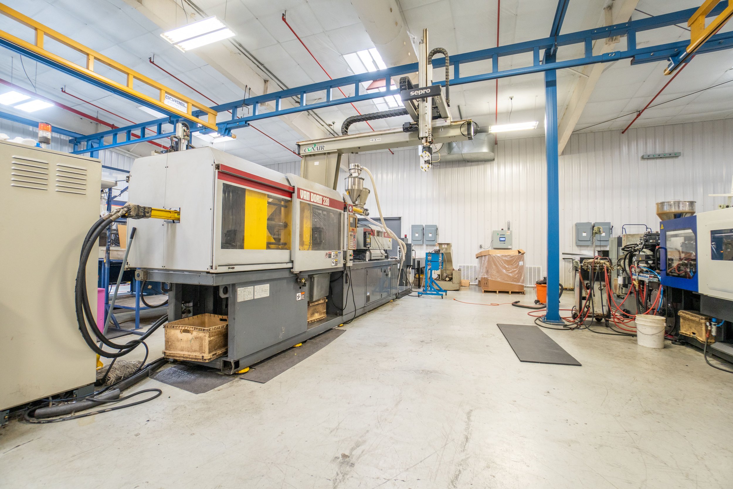 Industrial manufacturing room with large machinery, including a Van Doren 230 injection molding machine, electrical panels, and an overhead crane system.