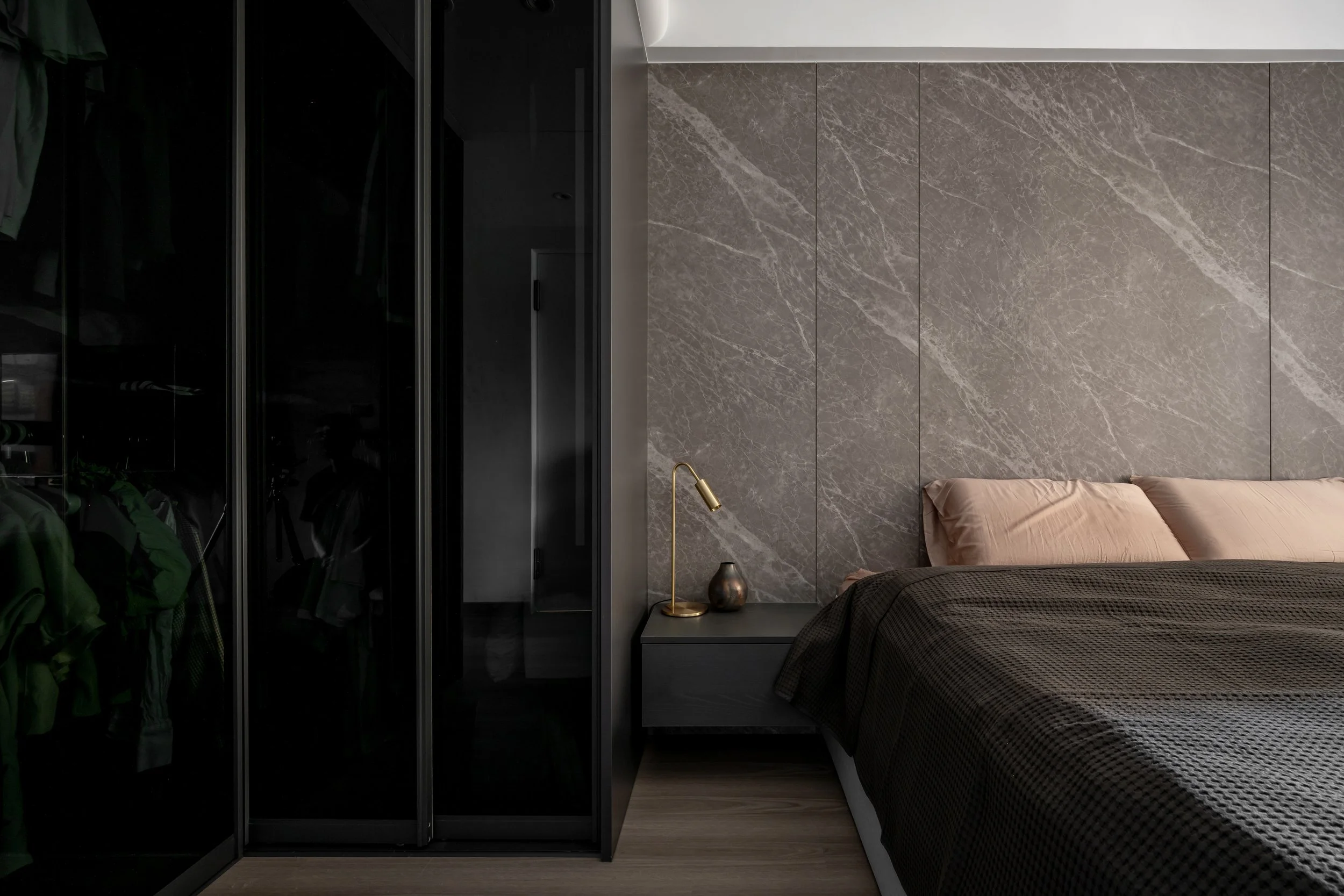 Modern bedroom with a bed, beige pillows, black and brown bedding, a gray marble accent wall, a black nightstand with a gold desk lamp and a metallic vase, and a closet with black glass doors.