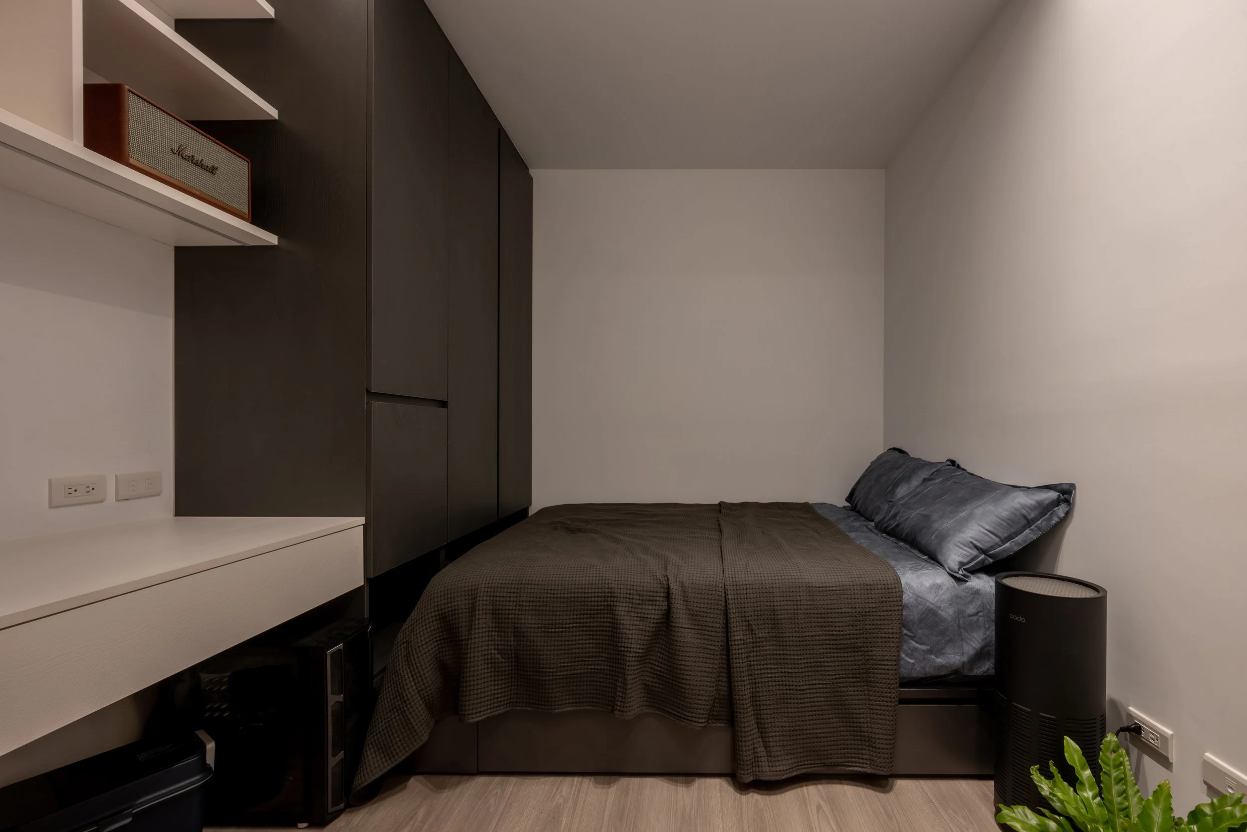 A small bedroom with a bed, dark bedding, and a black air purifier next to a white wall.