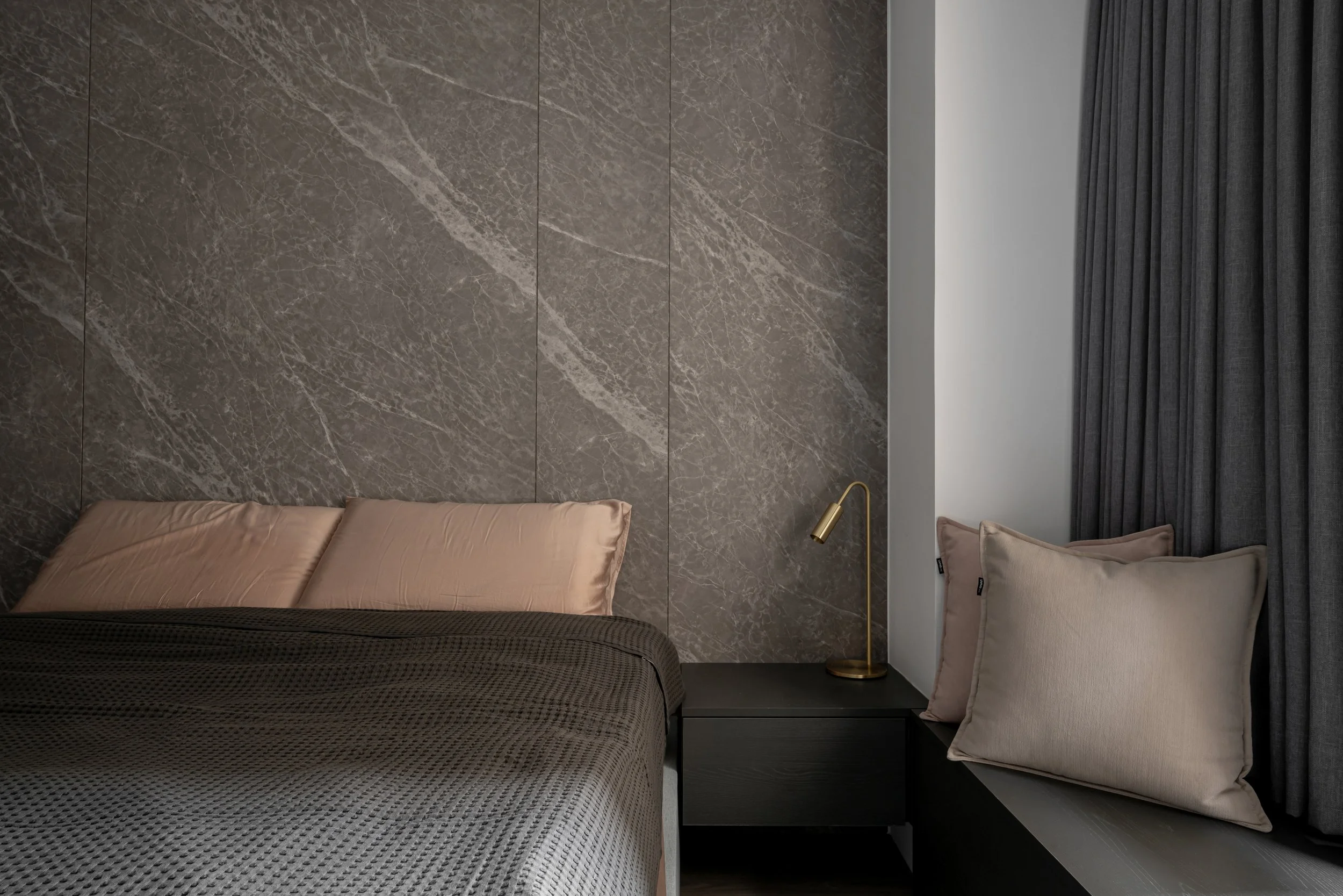 A modern bedroom corner with a gray marble wall, a black nightstand, a gold desk lamp, a beige pillow, beige curtains, and part of a bed with gray and beige bedding.