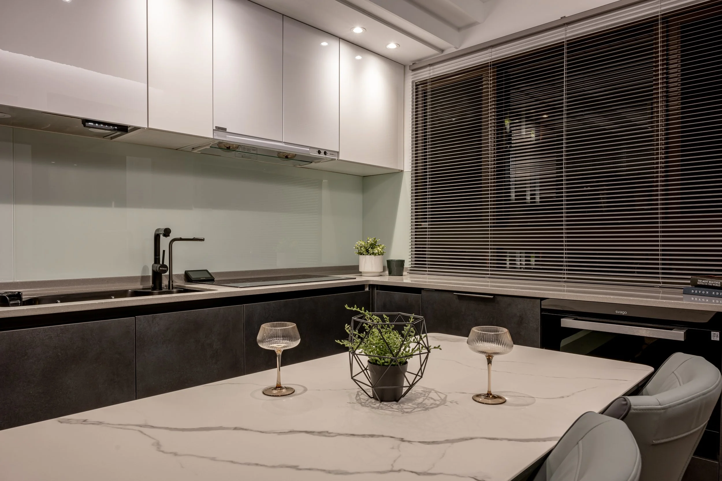Modern kitchen with white and black cabinetry, a marble dining table with two wine glasses and a potted plant, blinds covering a large window, and minimal decor.
