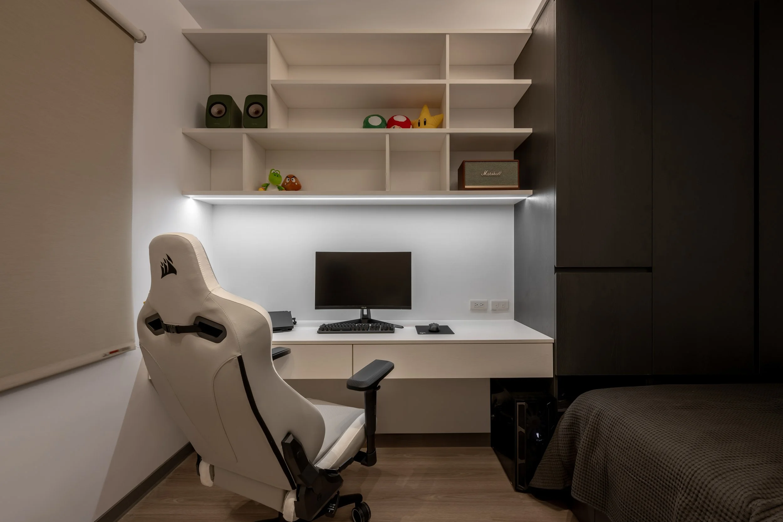 Minimalist workspace with a white desk, a computer monitor, a beige ergonomic chair, and black storage cabinets. Open shelves above the desk display toys and a grey speaker.
