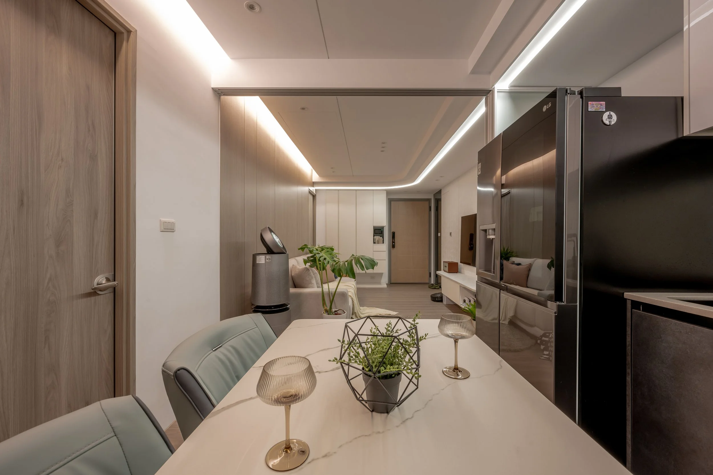 Modern living and dining area with a white marble table, decorative plants, and a large refrigerator in an open-plan apartment.