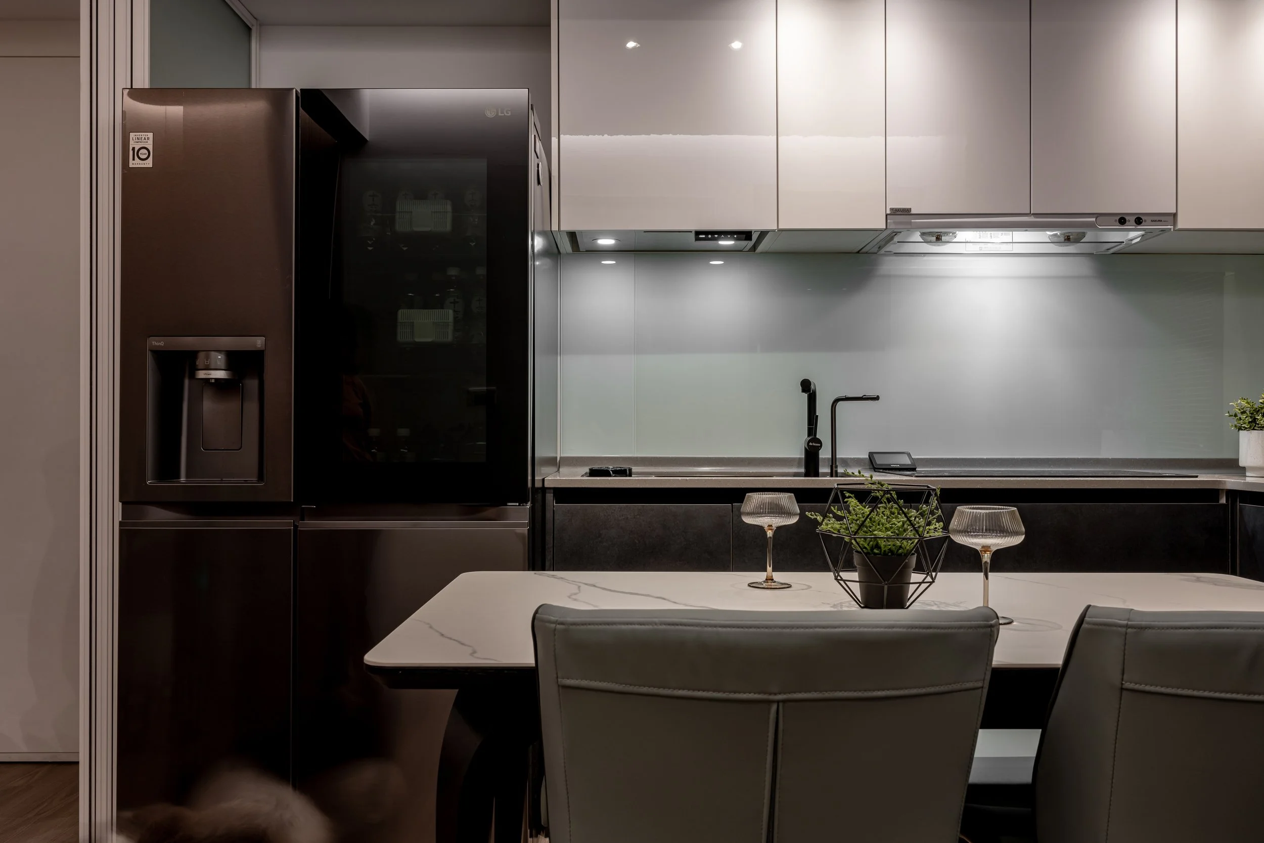 Modern kitchen with black refrigerator, white cabinets, gray countertop, black faucet, and dining table with two glasses, a plant in a wire basket, and gray chairs.