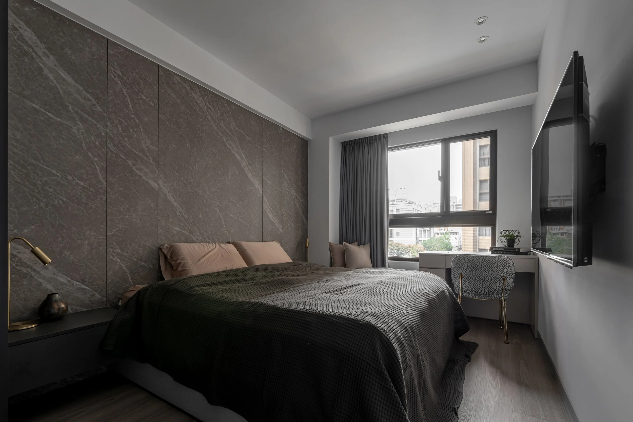 Modern bedroom with a large bed, dark bedding, and pink pillows, a textured accent wall behind the bed, a window with gray curtains, a white desk with a patterned chair, and a wall-mounted television.